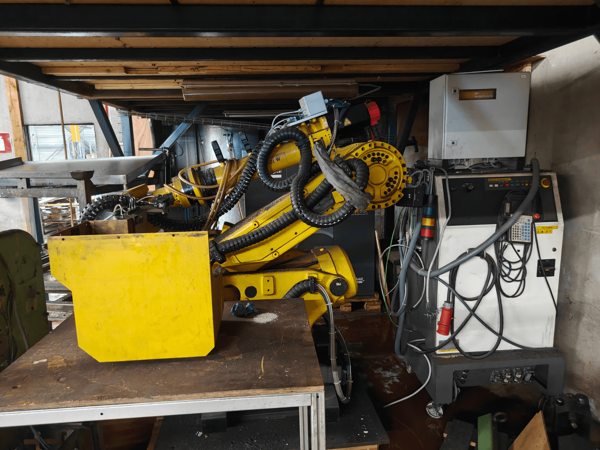 Vorderansicht eines FANUC-Roboterarms vom Typ S-430i W, gelb, mit Bedienfeld, Kabeln und einem gelben Bauteil auf einer Werkbank.