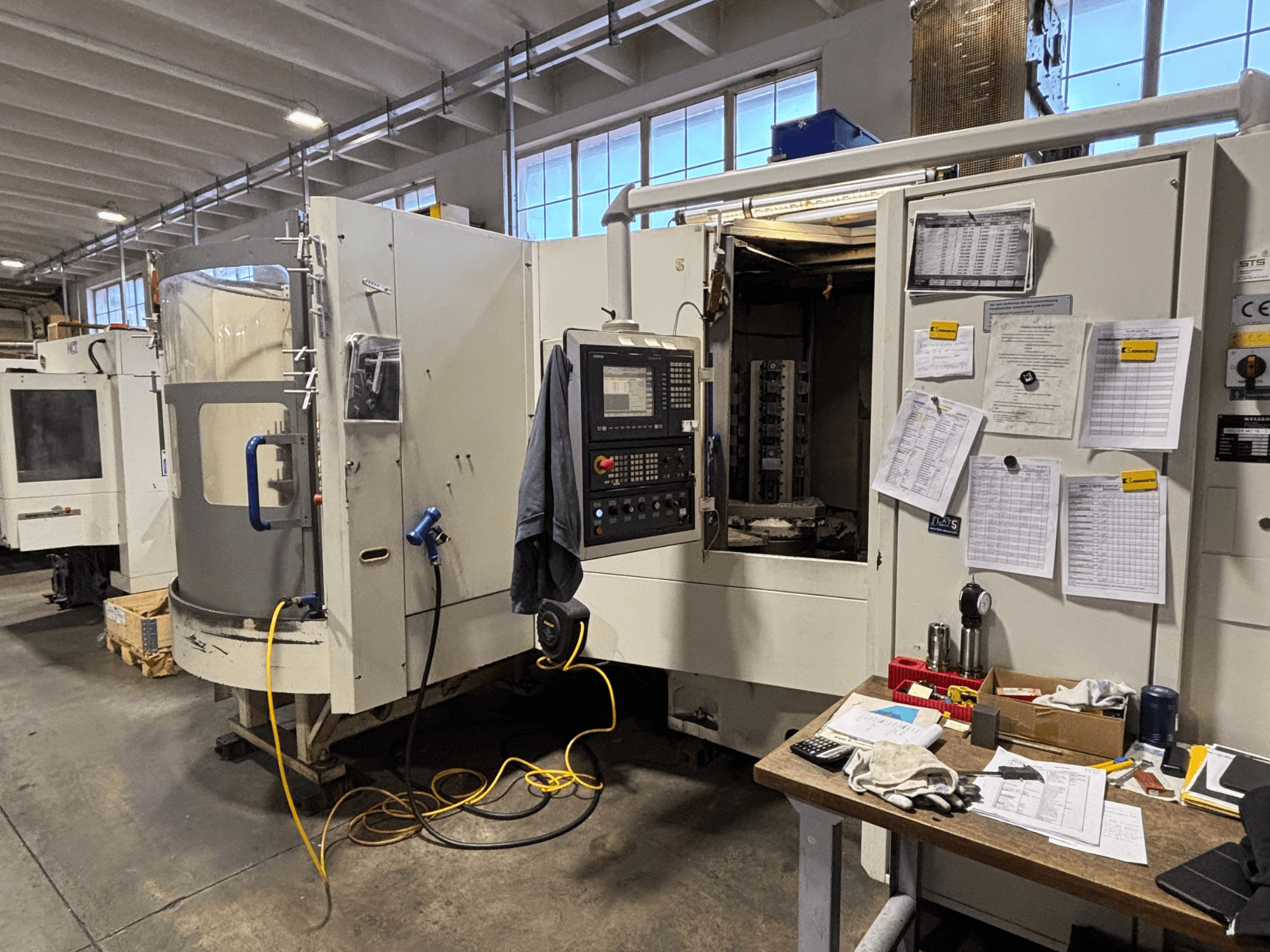 CNC-Maschine in einer Werkstatt, Vorderansicht, mit Bedienfeld und Dokumentation an der Seite; Werkzeuge und Materialien auf dem Tisch.