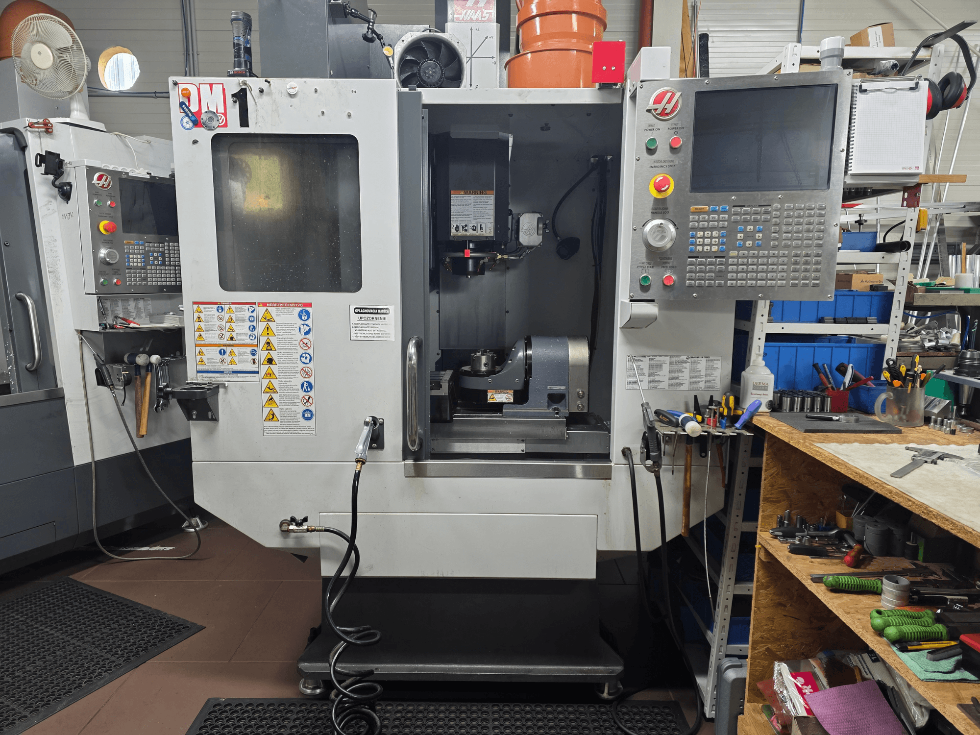 Haas CNC-Maschine Modell VF-2, Vorderansicht, mit Bedienelementen, Spindelbereich, Sicherheitsschildern und Werkzeugen auf einer Werkbank in der Nähe.
