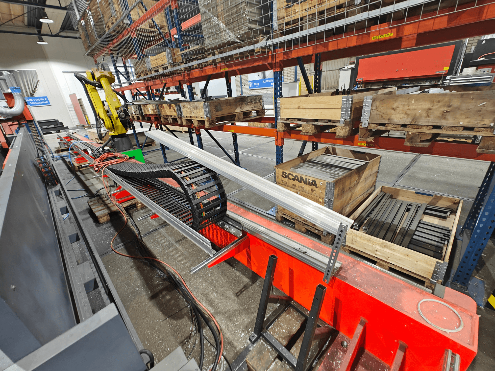 Roboterarm an einer Arbeitsstation, orangefarbene und schwarze Kabel sichtbar, mit Scania-Kisten und Aluminiumprofilen in einer Fabrikumgebung.