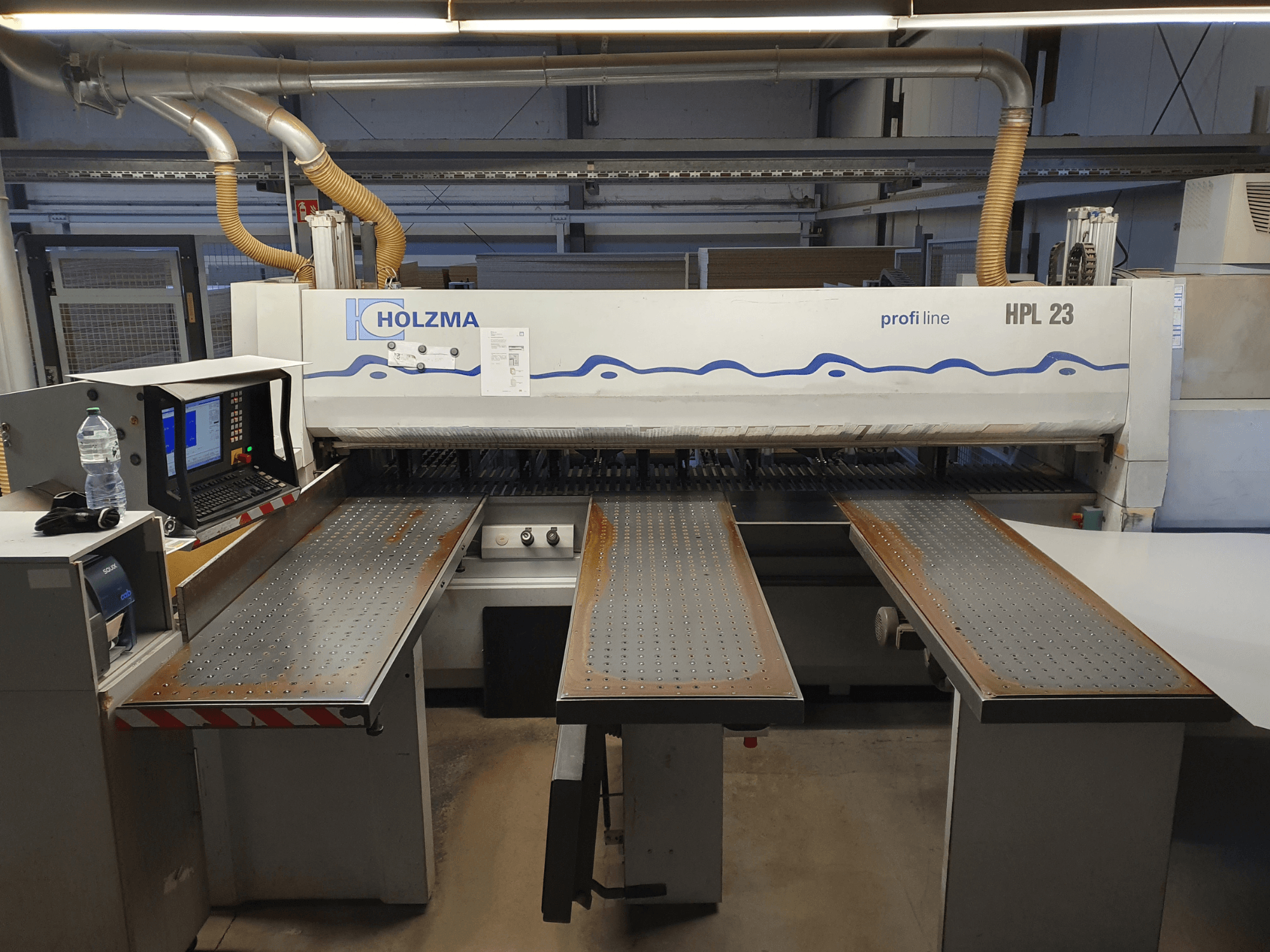 HOLZMA HPL 23 Schneidemaschine in der Vorderansicht, mit drei perforierten Tischen, Computerterminal und Wasserflasche daneben.