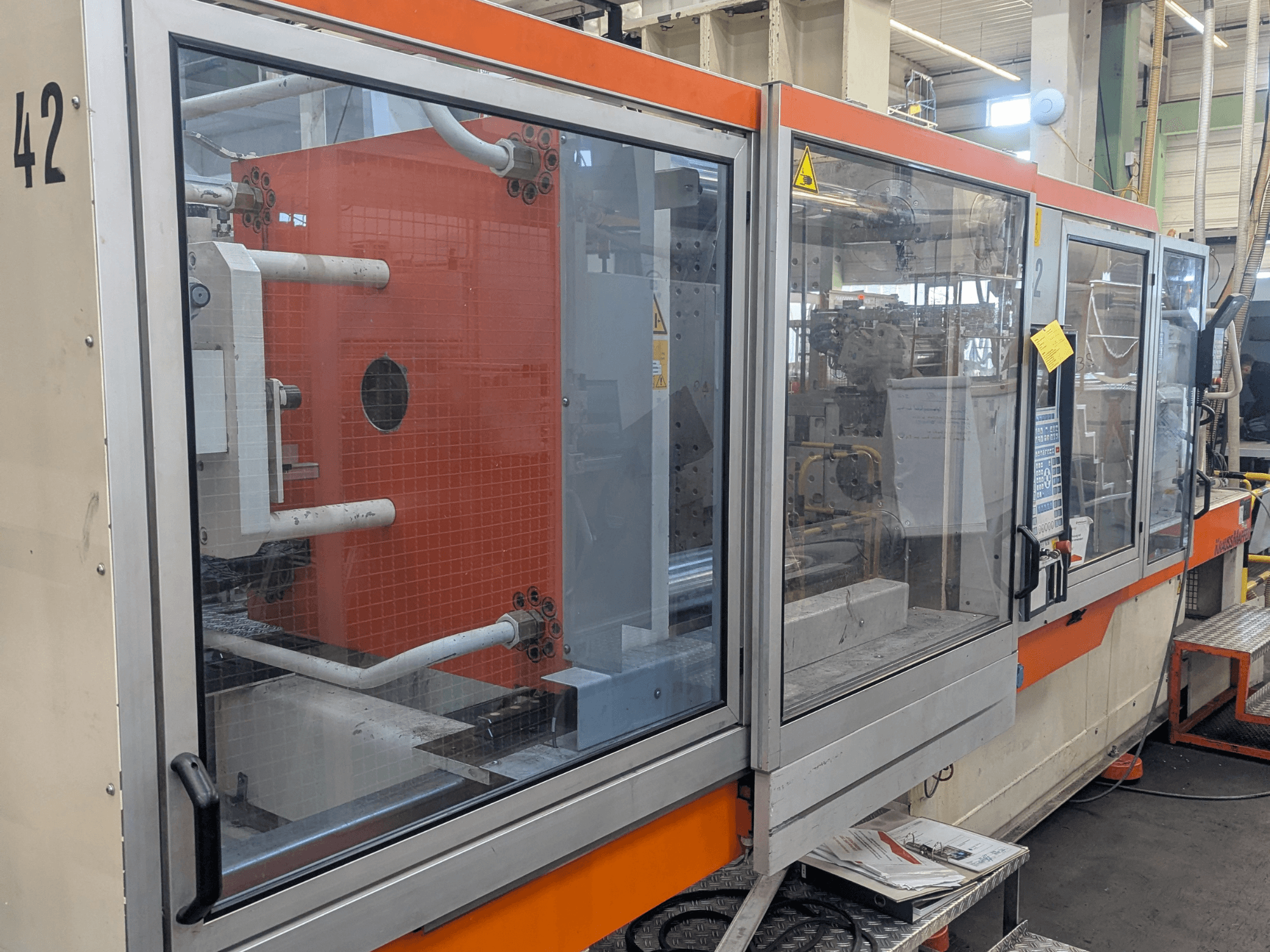 Hydraulische Spritzgießmaschine Krauss Maffei KM 350-3000, Seitenansicht mit Bedienelementen und internen Komponenten hinter Glasscheibe.