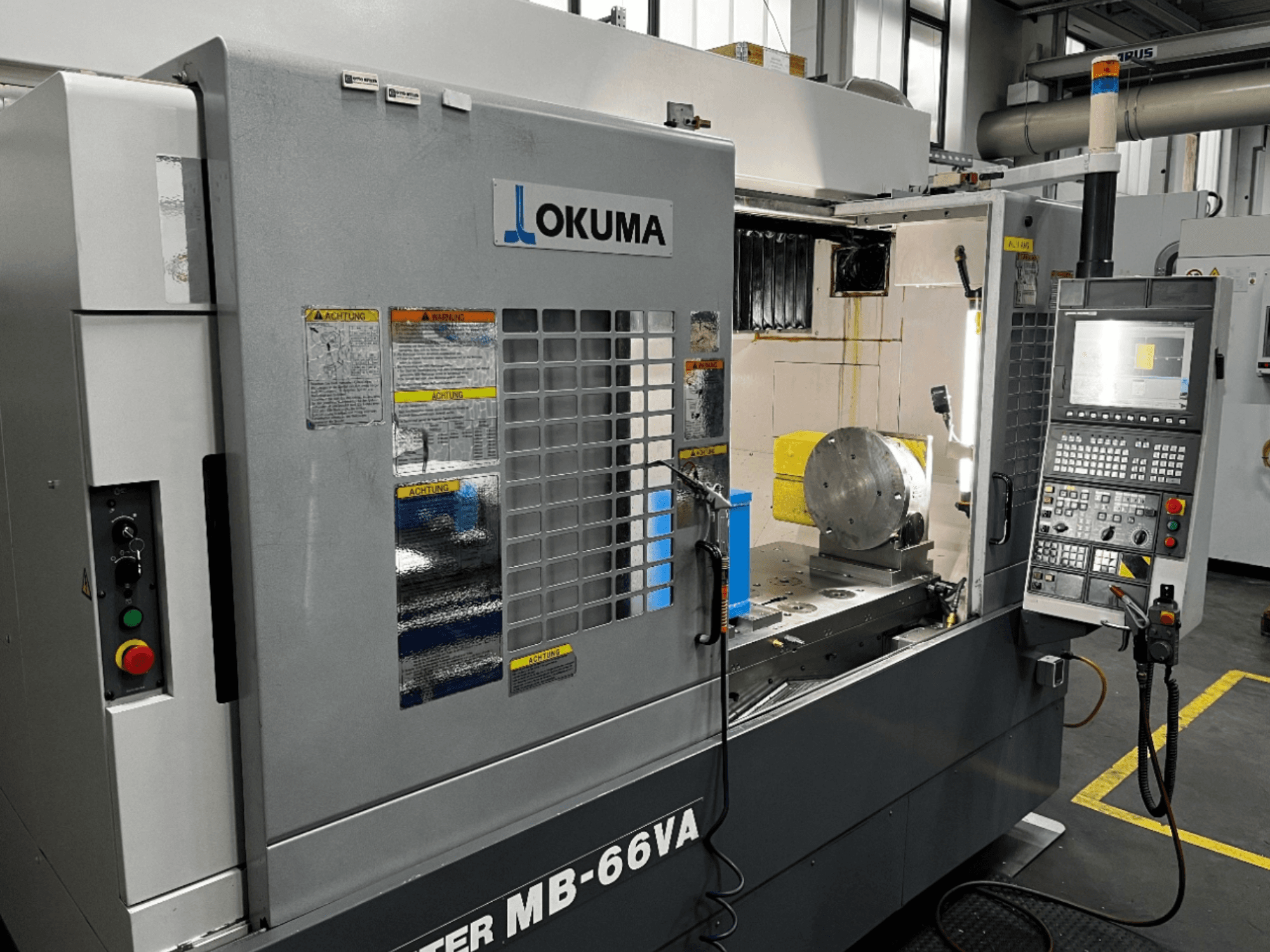 Okuma MB66-VA Vertikal-Bearbeitungszentrum in der Frontansicht mit Bedienfeld, Werkstück und Maschineninnenraum mit Sicherheitsschildern.