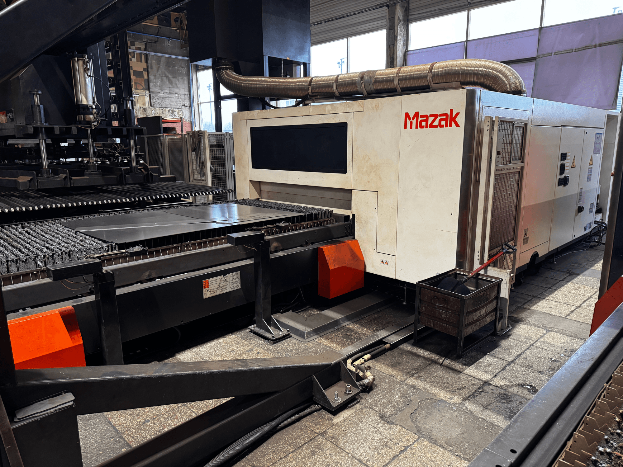 Mazak-Laserschneidmaschine in einer Werkstatt, Seitenansicht mit Bedienfeld, Förderband und Auffangbehälter.