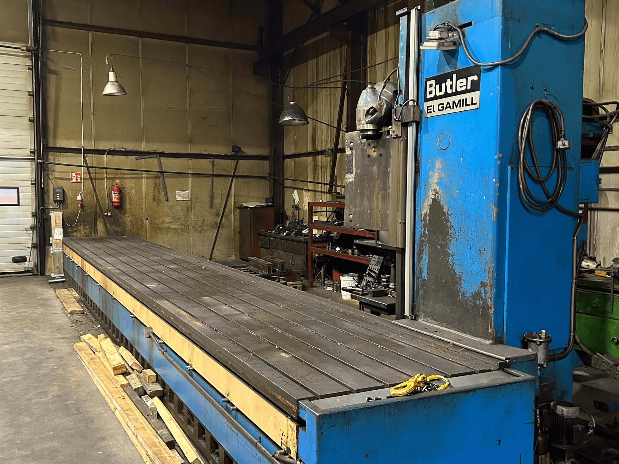 Blaue Butler-El-Gamill-Fräsmaschine in einer Werkstatt, Seitenansicht mit langem, flachem Tisch und Bedienelementen auf der rechten Seite.