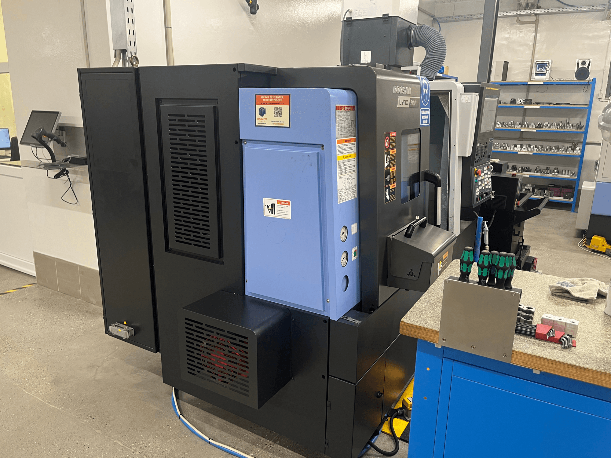 Doosan Lynx 2100 Drehmaschine, Seitenansicht, mit Bedienfeld, Belüftungssystem und Werkzeugen auf einer Werkbank.