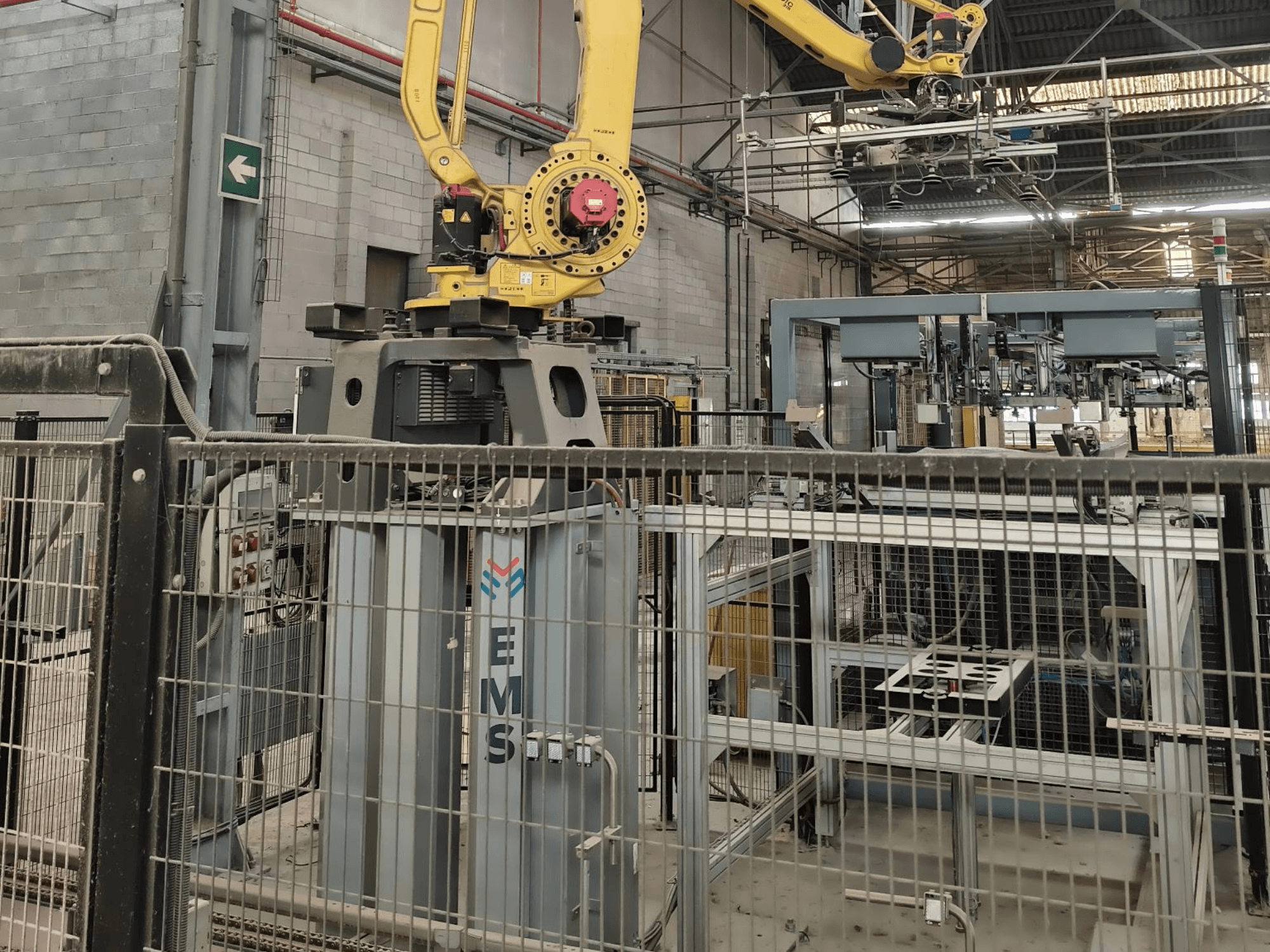 Vorderansicht des FANUC M-410iC/315-Roboterarms, der über einem EMS-Werkzeug positioniert ist, im Hintergrund sind Industrieanlagen und eine Umzäunung zu sehen.