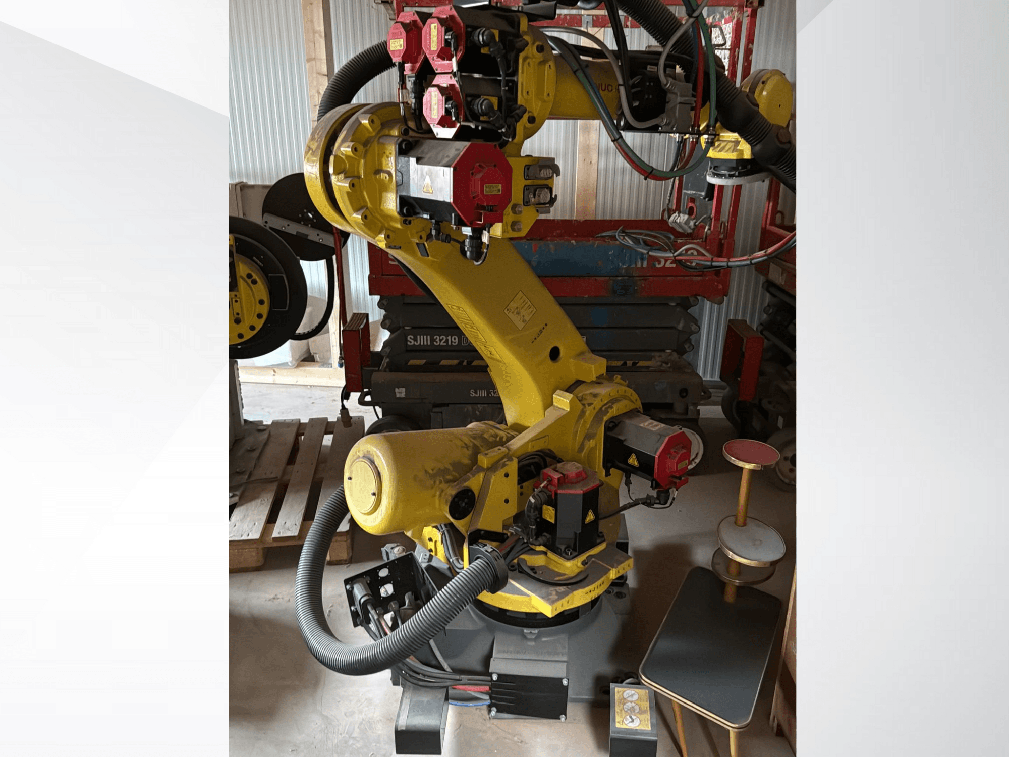 Gelber Industrieroboterarm mit roten Steuergeräten, Vorderansicht; mit mehreren Gelenken und Schläuchen, auf einer Baustelle aufgestellt.