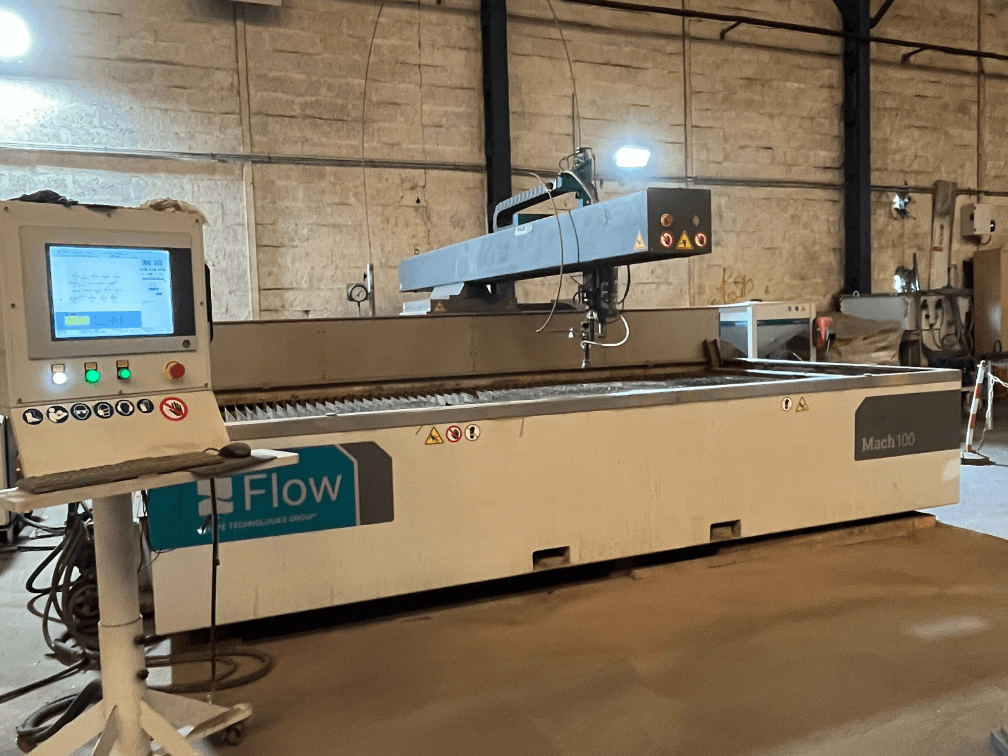 Flow Mach 100 Wasserstrahlschneider, Seitenansicht, mit digitalem Bedienfeld und Schneidkopf auf einem Arbeitstisch.