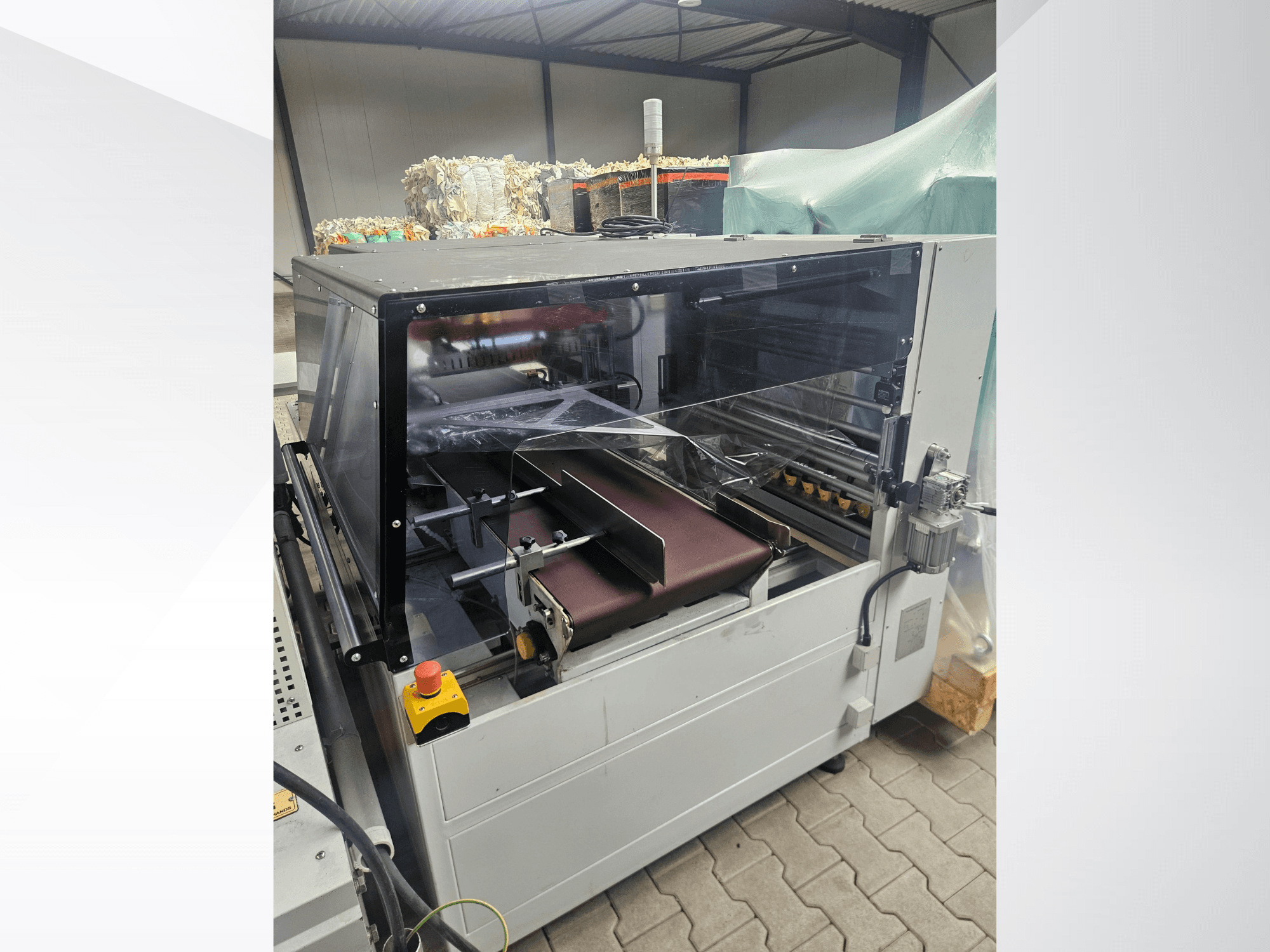 AMTEC Speed Shrink 40 PRO-Maschine, Vorderansicht, mit einem Kunststoffverarbeitungsmechanismus mit durchsichtiger Schutzabdeckung und Förderband.