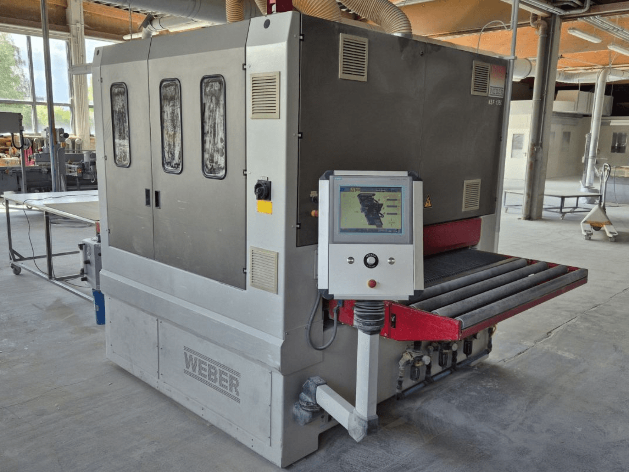 WEBER KSF-3-1350 Bandschleifmaschine, Seitenansicht mit Bedienelementen, Bedienfeld und Schleifband. Industrielle Umgebung mit sichtbaren Maschinen.
