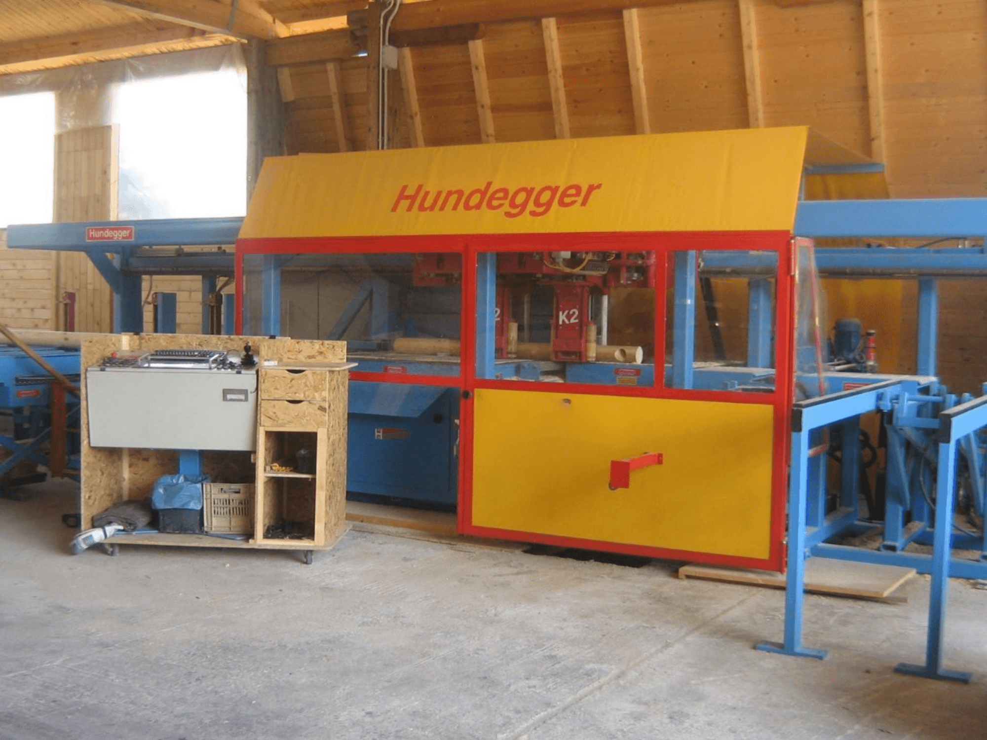 CNC-Holzbearbeitungszentrum HUNDEGGER K2 4 assi in einer Werkstatt, Vorderansicht mit gelb-blauem Design und Arbeitstisch.