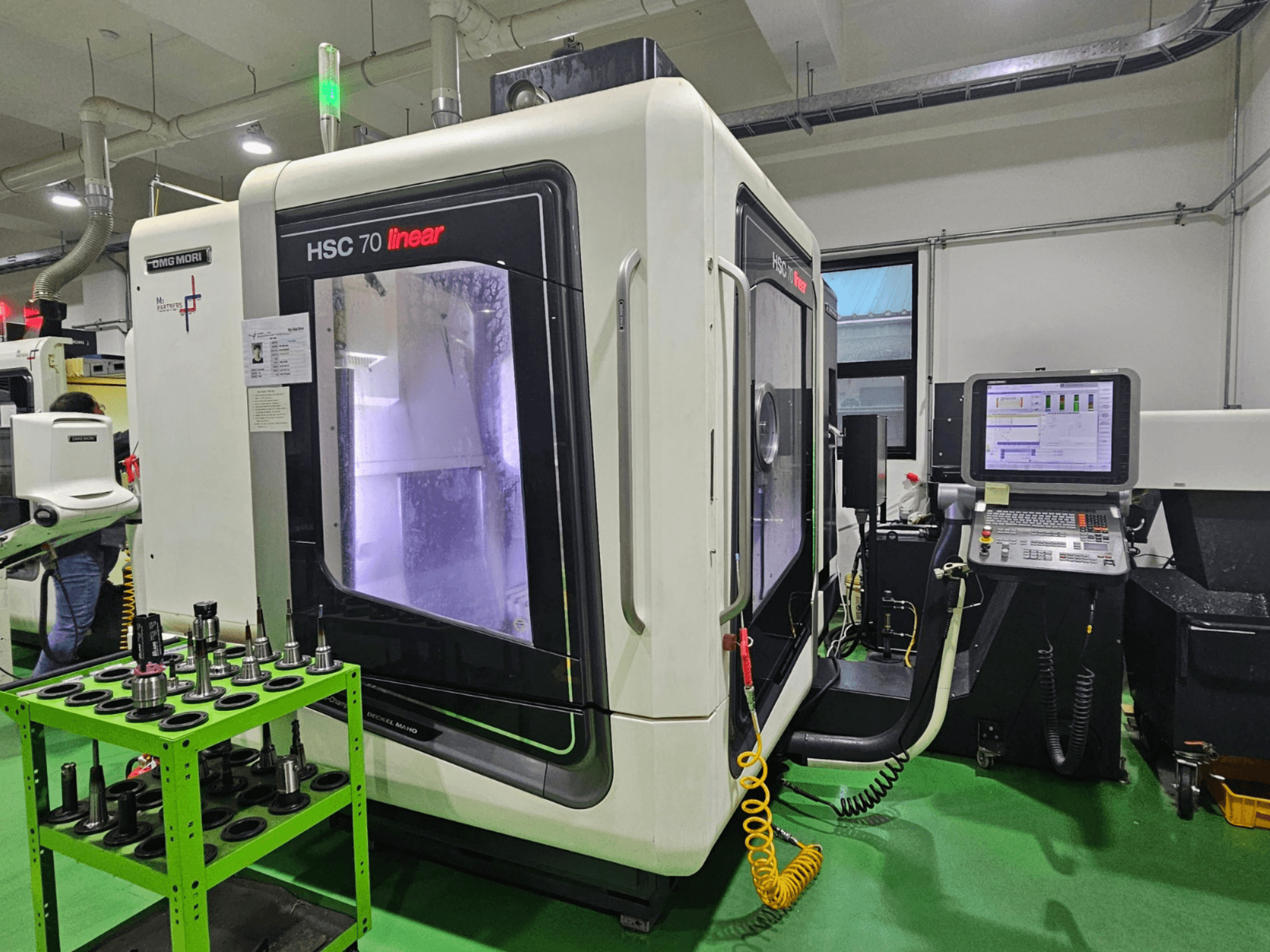 DMG MORI HSC 70 Linear-Universal-Bearbeitungszentrum, Vorderansicht, mit Werkzeughaltern auf einem grünen Tisch und einem sichtbaren Bedienfeld.