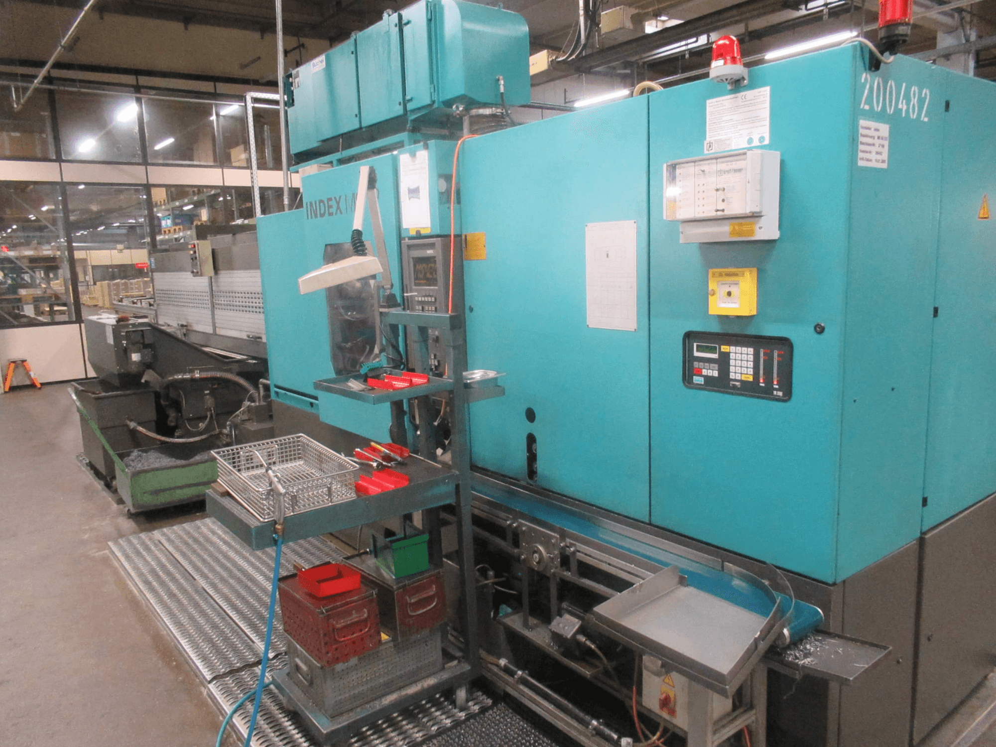 Türkisfarbene INDEX-Maschine in der Fabrik, Seitenansicht; mit Bedienfeld, Werkzeugspeicher und Förderband. Sichtbare Fächer, Schläuche und Alarme.