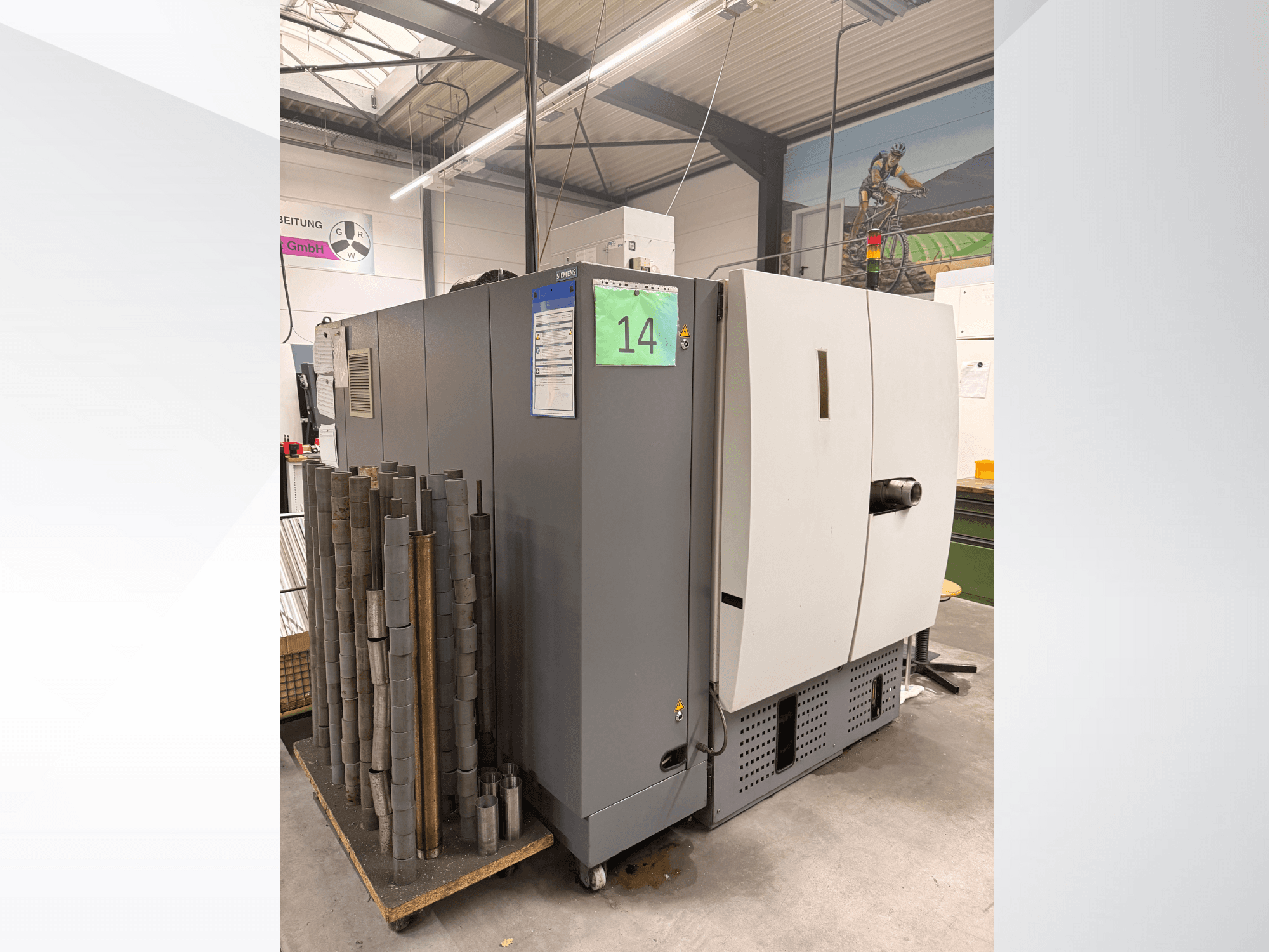 Graue Industriemaschine von Siemens, Seitenansicht, mit einem Lager zylindrischer Metallteile daneben und sichtbaren Geräteetiketten.