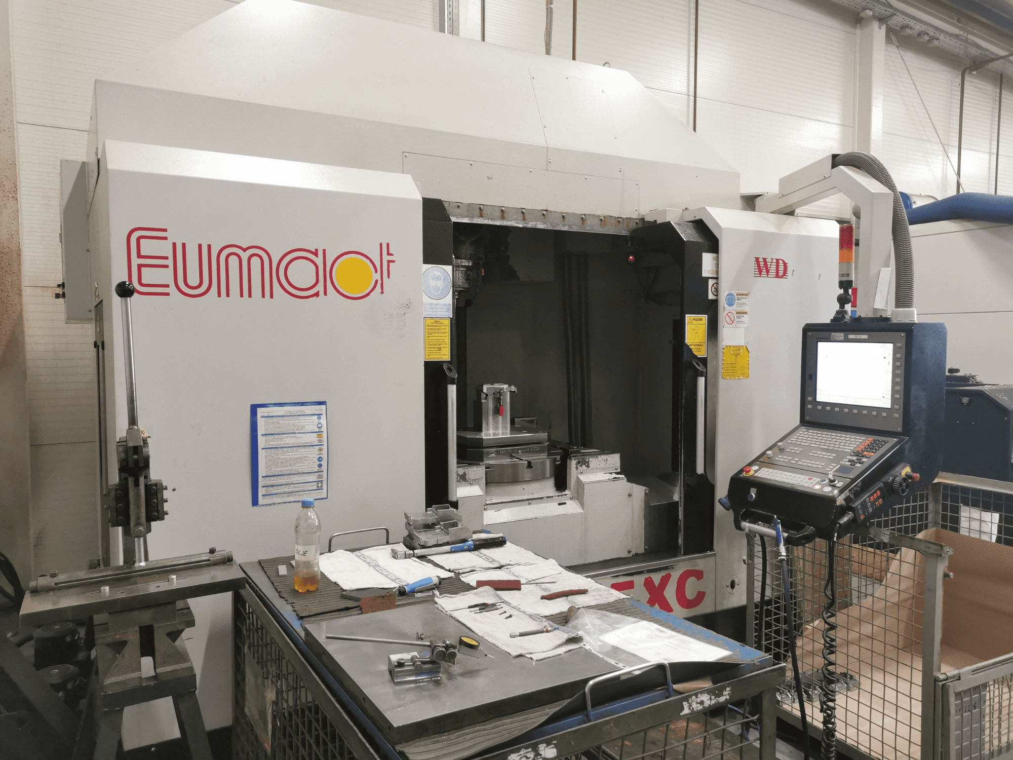Vorderansicht einer Eumdaot-CNC-Maschine mit Bedienfeld, Werkzeugen und einer mit Bauteilen und Materialien vollgestopften Arbeitsfläche.