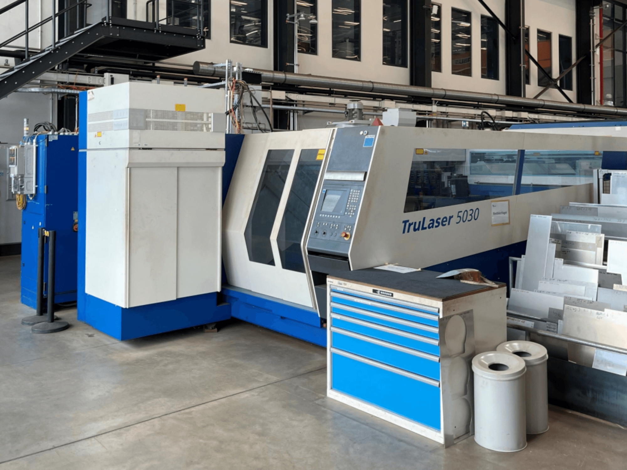 TRUMPF TruLaser 5030 CO₂-Laserschneidmaschine in der Vorderansicht, mit Bedienfeld und angrenzender Werkbank mit Blechen.