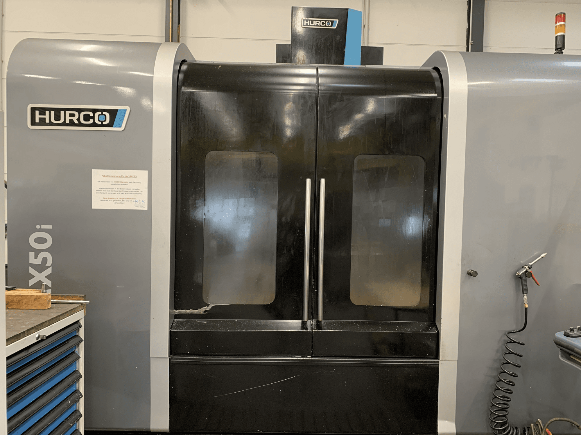 Vorderansicht einer Hurco VX50i CNC-Maschine mit Doppeltüren, mit technischen Bedienelementen und einem Wartungsschild an der Seite.