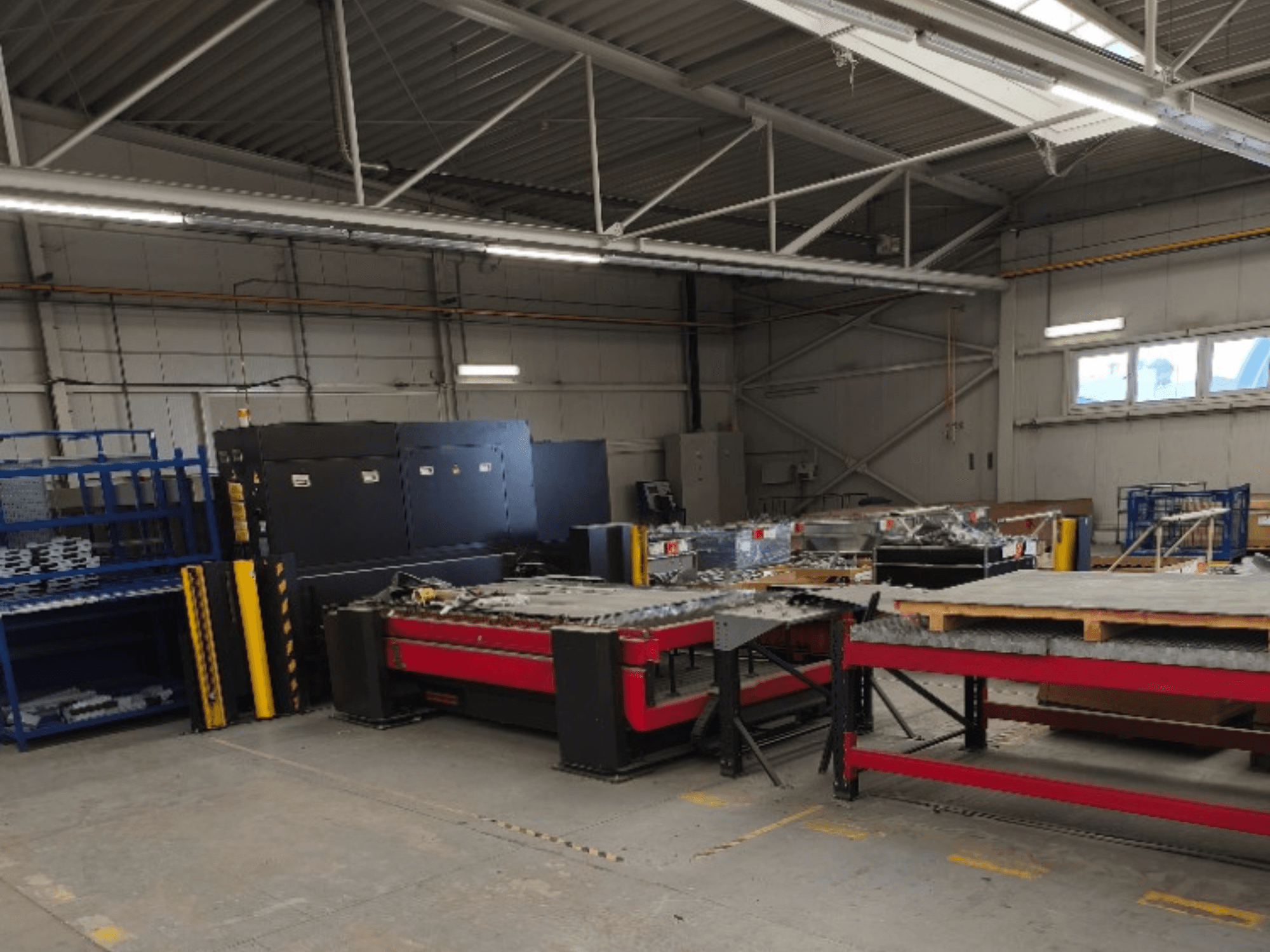 Eine AMADA LCG 3015 AJ Faserlaserschneidemaschine in einer Werkstatt, von vorne gesehen, mit Lagerregalen und Materialien im Hintergrund.