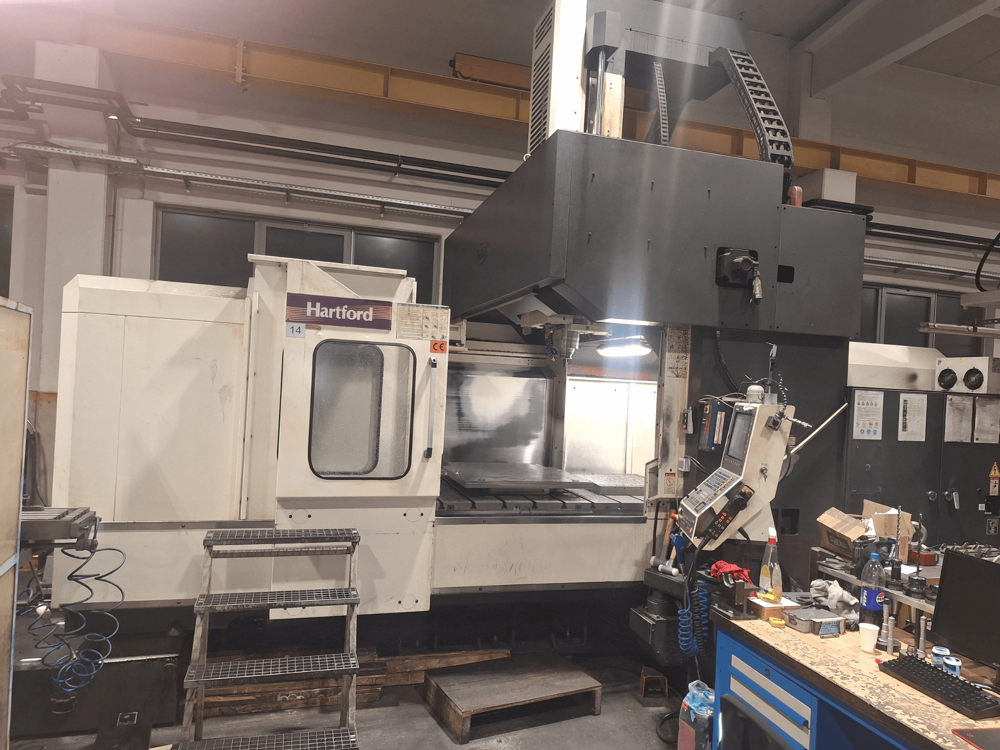 Hartford-CNC-Maschine, Vorderansicht, mit technischen Bedienelementen, Zugangstreppe und einem Arbeitstisch in einer Werkstattumgebung.