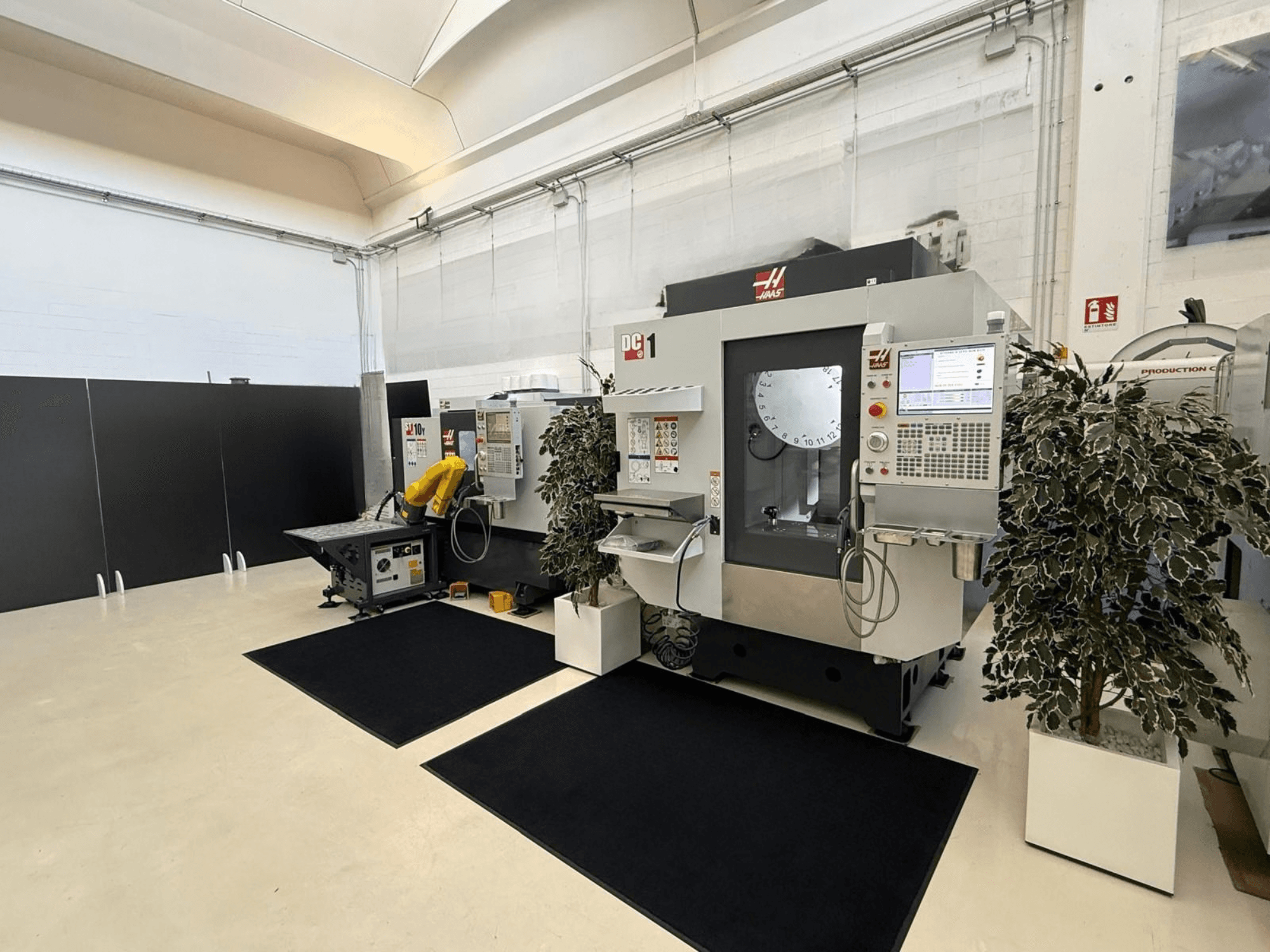 Vorderansicht eines vertikalen Bearbeitungszentrums vom Typ HAAS DC-1 in einem sauberen Arbeitsbereich, mit einem Touchscreen-Bedienfeld und den umliegenden Anlagen.