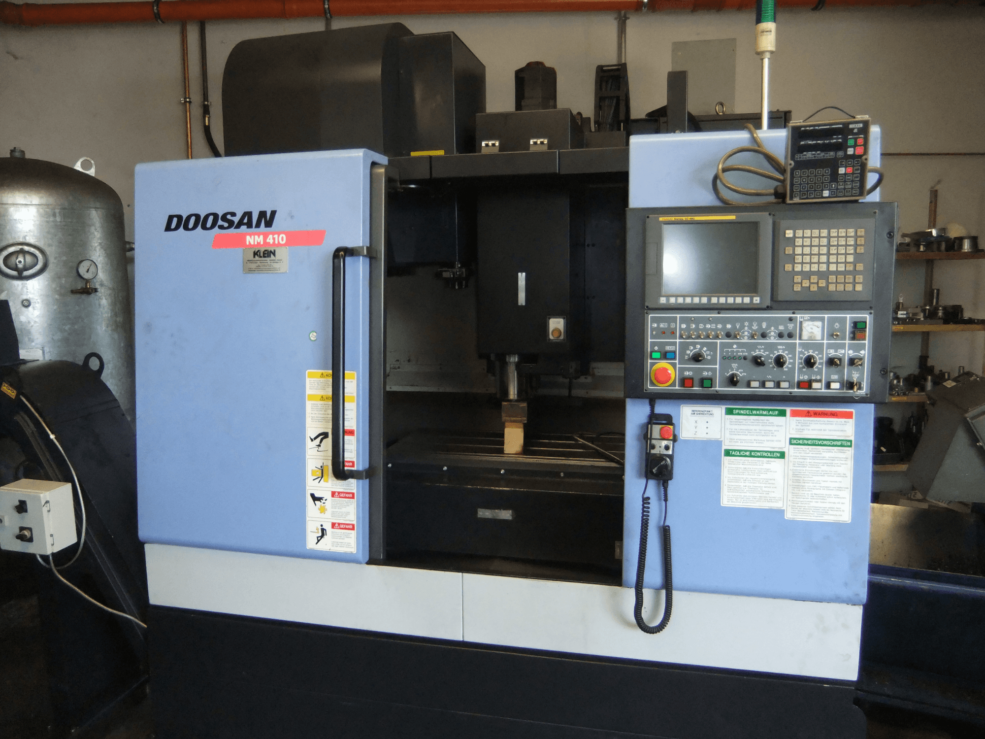 Vertikales Bearbeitungszentrum DOOSAN NM 410, Vorderansicht, mit Blick auf das Bedienfeld, den Spindelbereich und die Sicherheitshinweise.