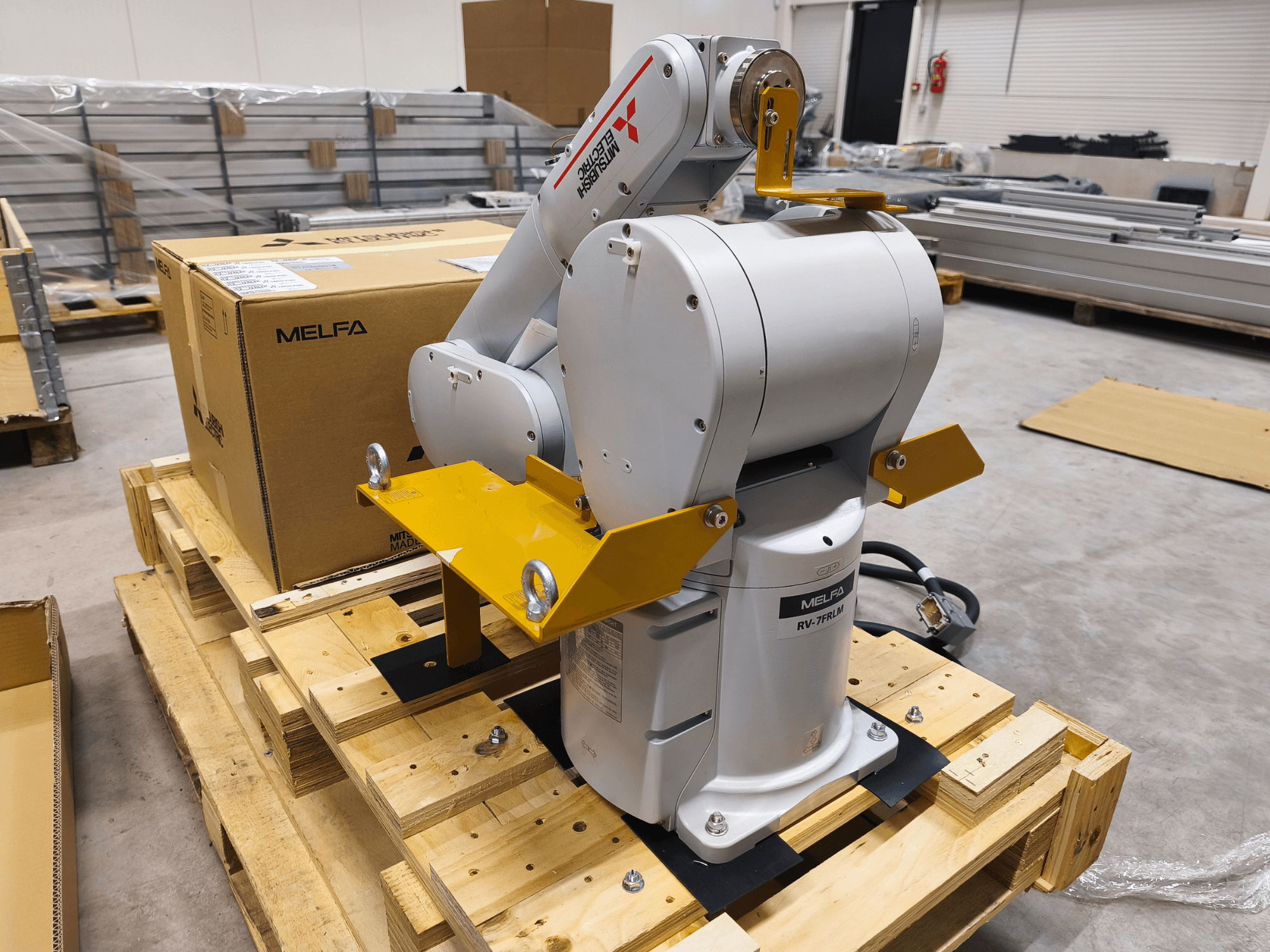 MELFA RV-7FLM Industrieroboterarm auf Holzpalette, Vorderansicht, mit gelben Halterungen und grauem Verpackungskarton.