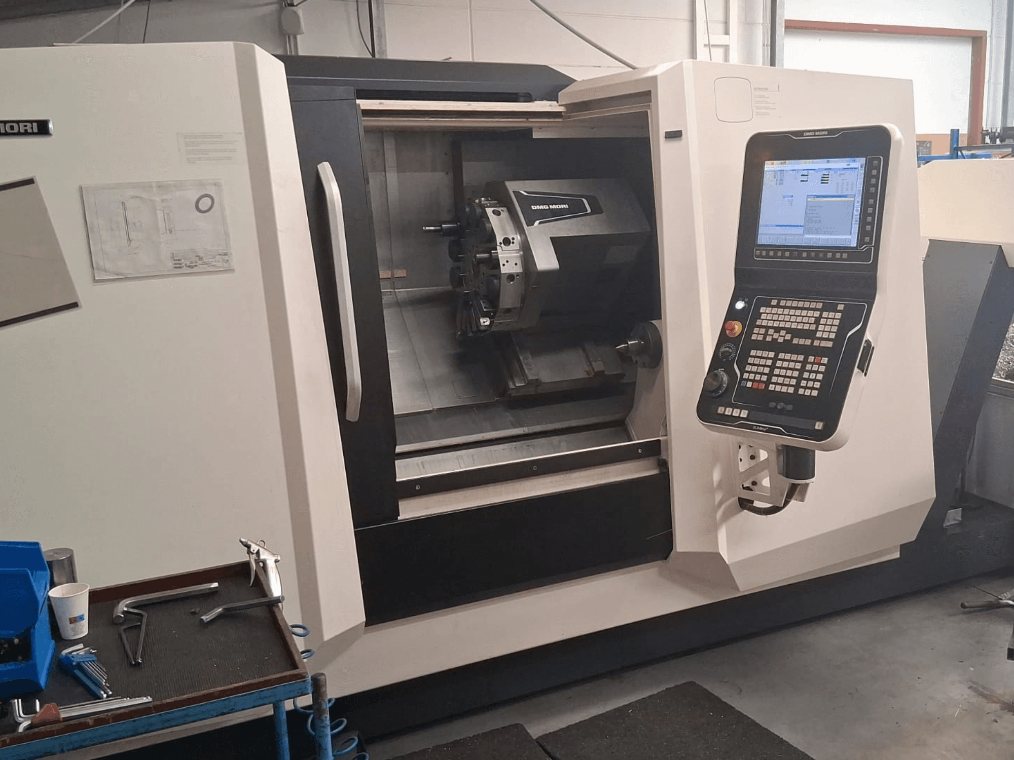 DMG MORI CLX450 Horizontaldrehmaschine in der Vorderansicht, mit Bedienfeld und Arbeitsbereich mit Werkzeugen und Dokumentation.