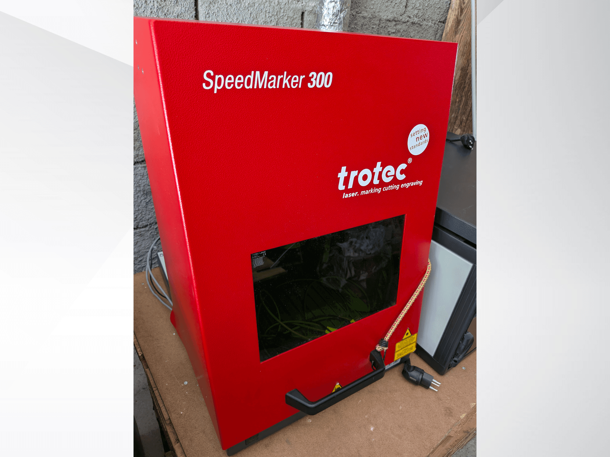 Rotes Trotec SpeedMarker 300 Laserbeschriftungsgerät in der Frontansicht, mit großem Sichtfenster und Stromanschluss.