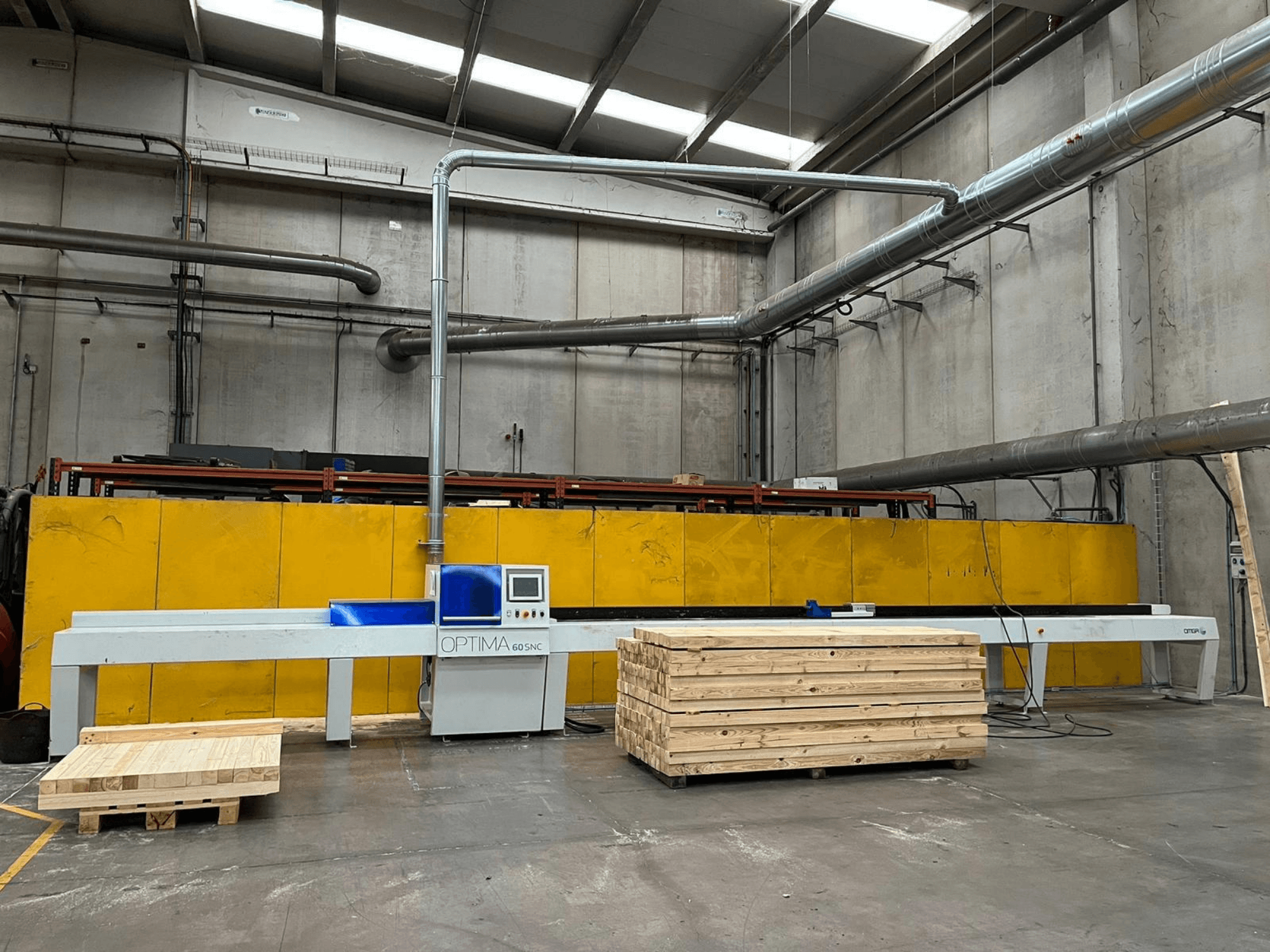 Vorderansicht der OPTIMA 60SNC 6200 CE-Optimierungs-Quersäge, im Hintergrund sind Holzpaletten und gelbe Sicherheitsbarrieren zu sehen.