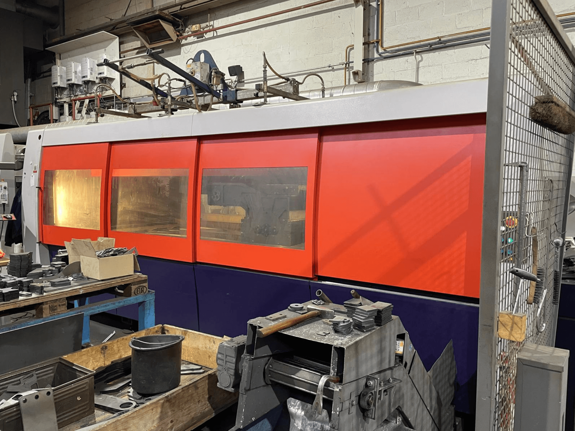 Nahaufnahme einer orange-weißen industriellen Laserschneidmaschine, Seitenansicht, mit Bedienfeld und Werkstattwerkzeugen im Vordergrund.