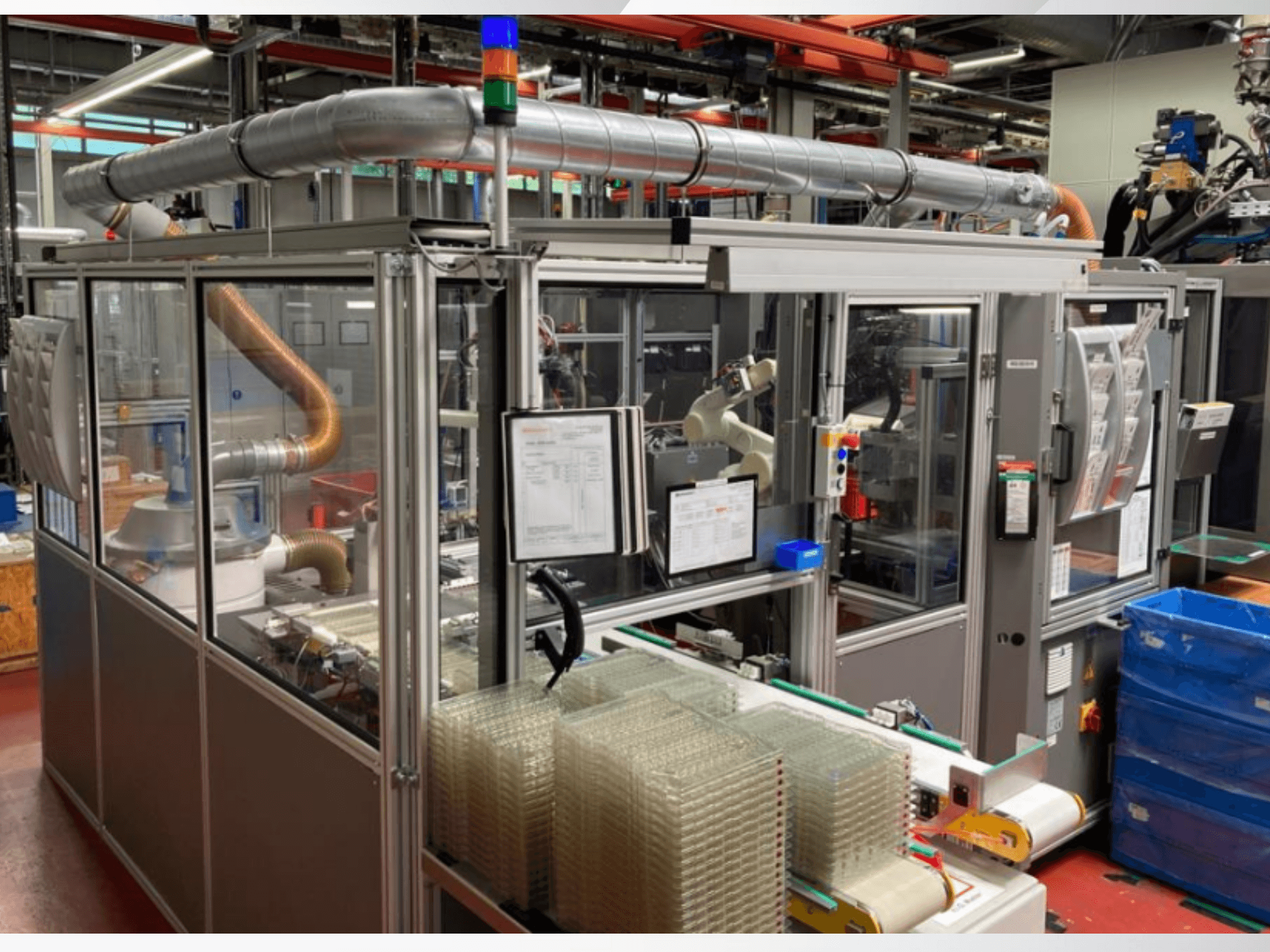 Vorderansicht einer automatisierten Maschine mit Metallrohren, Bedienfeld und gestapelten durchsichtigen Kunststoffbehältern in einer Fabrikhalle.