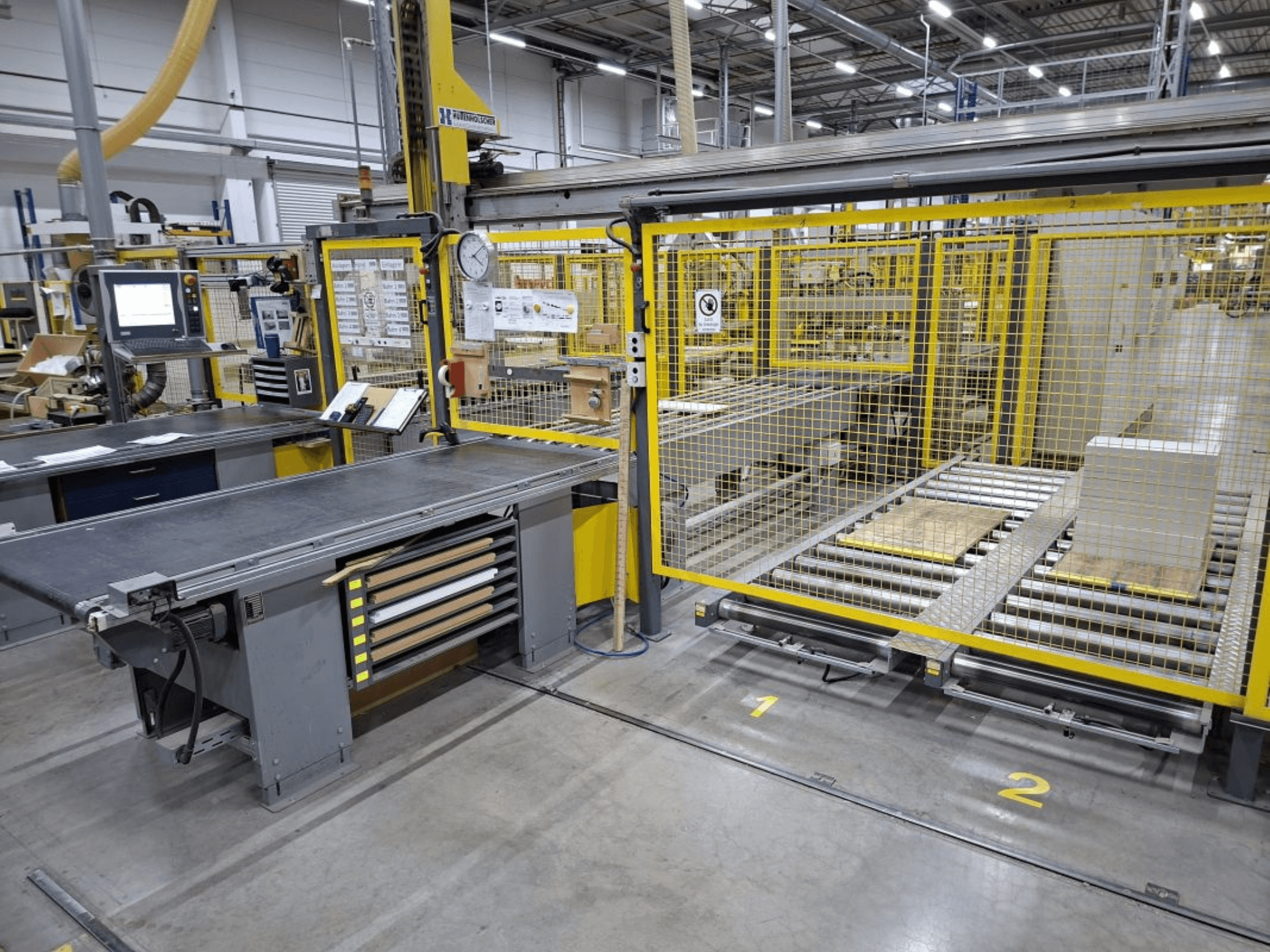 Industrieller Arbeitsbereich mit Förderanlage und Bedienfeld; Seitenansicht mit Maschinen und Schutzzaun.