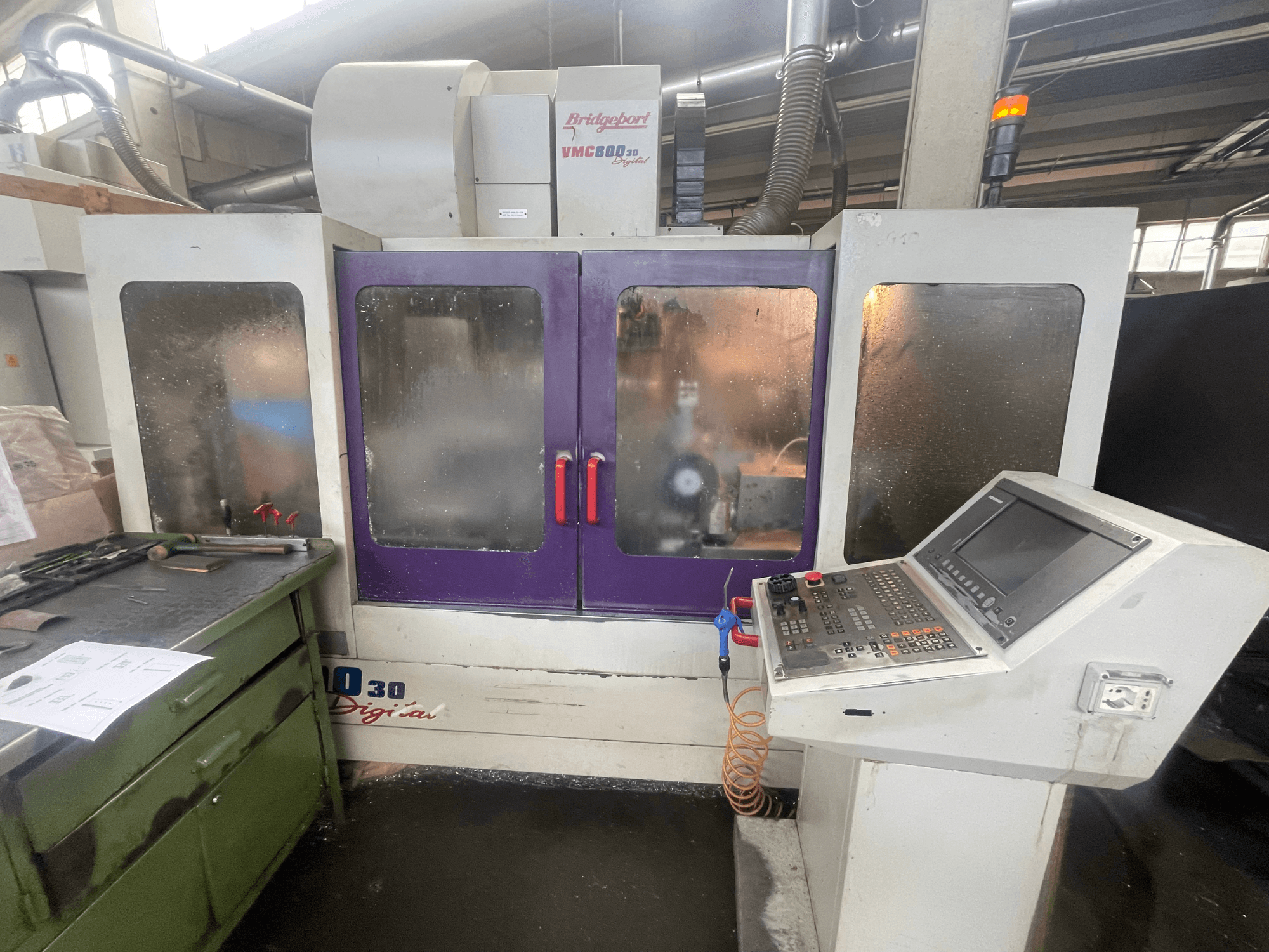 Vertikales Bearbeitungszentrum Bridgeport VMC 800/30, Vorderansicht, mit Bedienfeld und violetten Türen; Werkzeuge auf der angrenzenden Fläche.