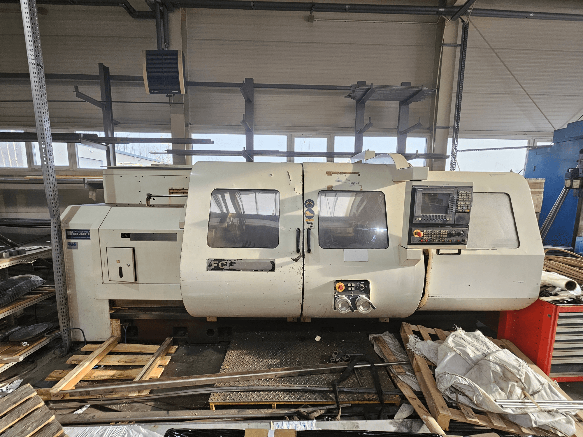 Wabeco CNC-Drehmaschine, Seitenansicht, mit Bedienfeld, Fenstern und Arbeitsbereich; Maschinen auf Holzpaletten in einer Werkstattumgebung.