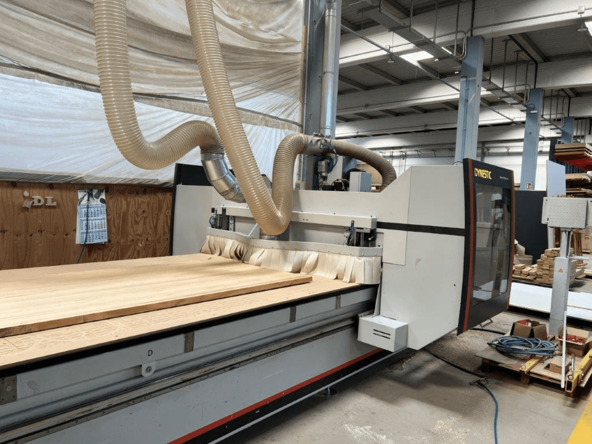 CNC-Maschine von Dynestic, Vorderansicht, mit einem großen Holzwerkstück und einem Absaugschlauch in einer Werkstatt.