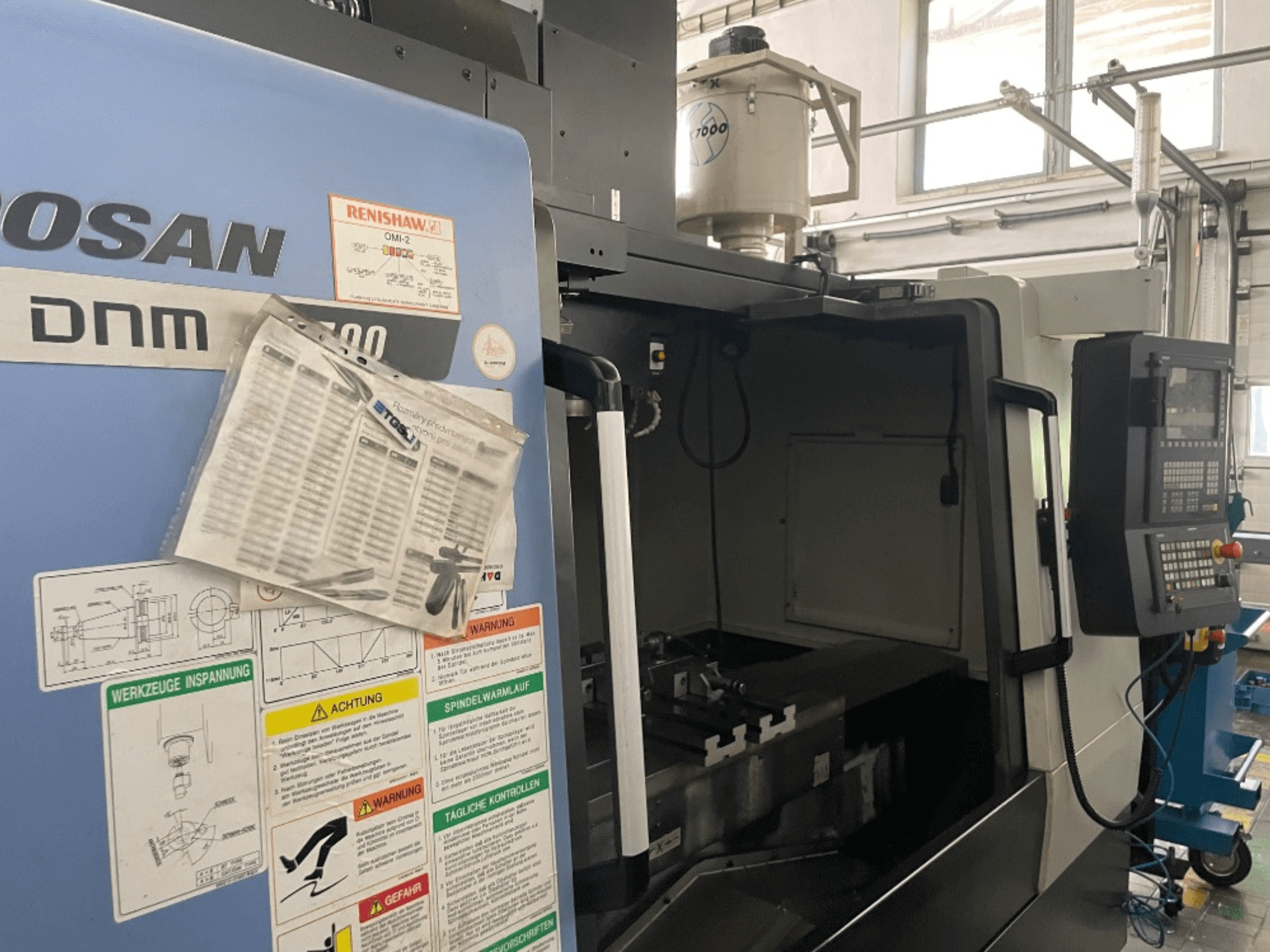 Vertikales Bearbeitungszentrum DOOSAN DNM 5700, Vorderansicht, zeigt Bedienfeld, Sicherheitsschilder und Maschinendetails.