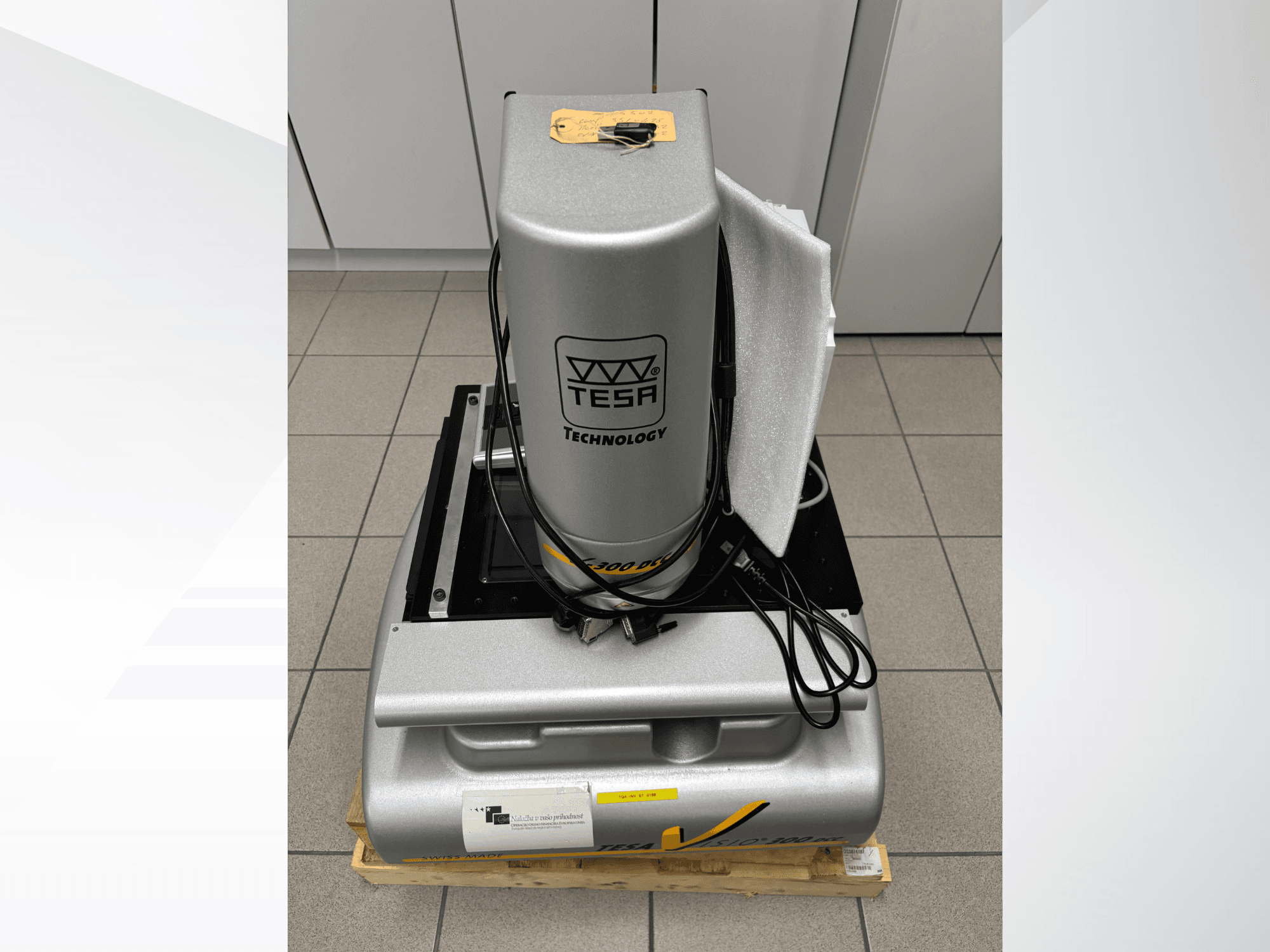 Optisches Messgerät TESA Visio 300 DCC in der Frontansicht mit Kabeln, auf einer Holzpalette, mit sichtbarer Schutzverpackung.