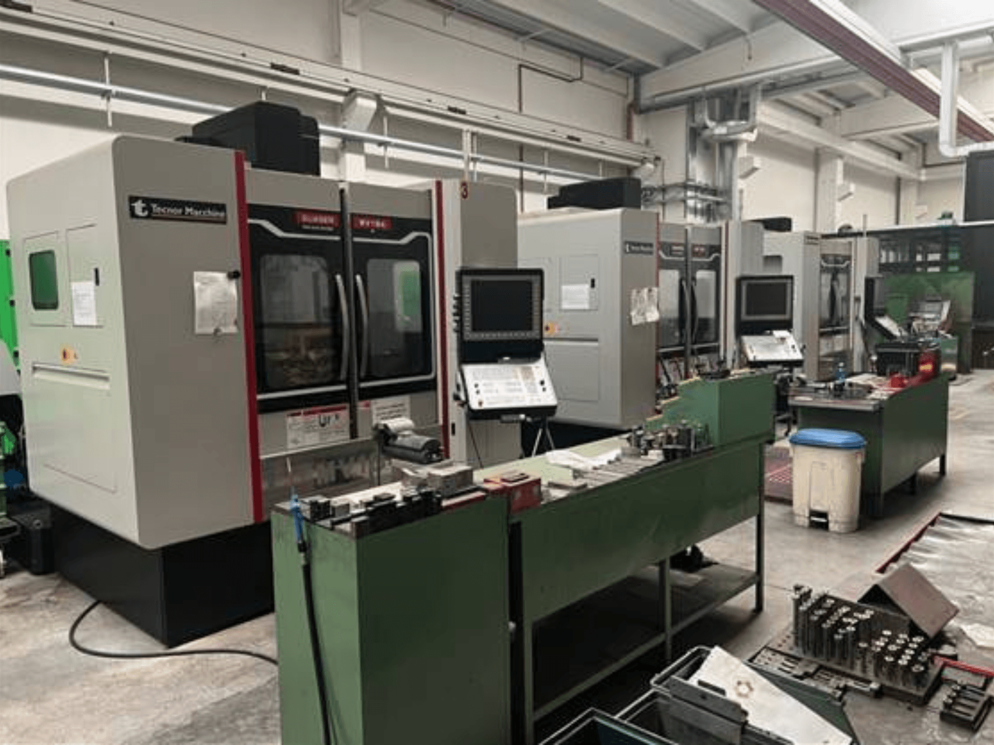 Vier CNC-Maschinen von Tecnoer Marchine in einer Werkstatt, Vorderansicht, mit Werkzeugen und Teilen auf grünen Werkbänken.