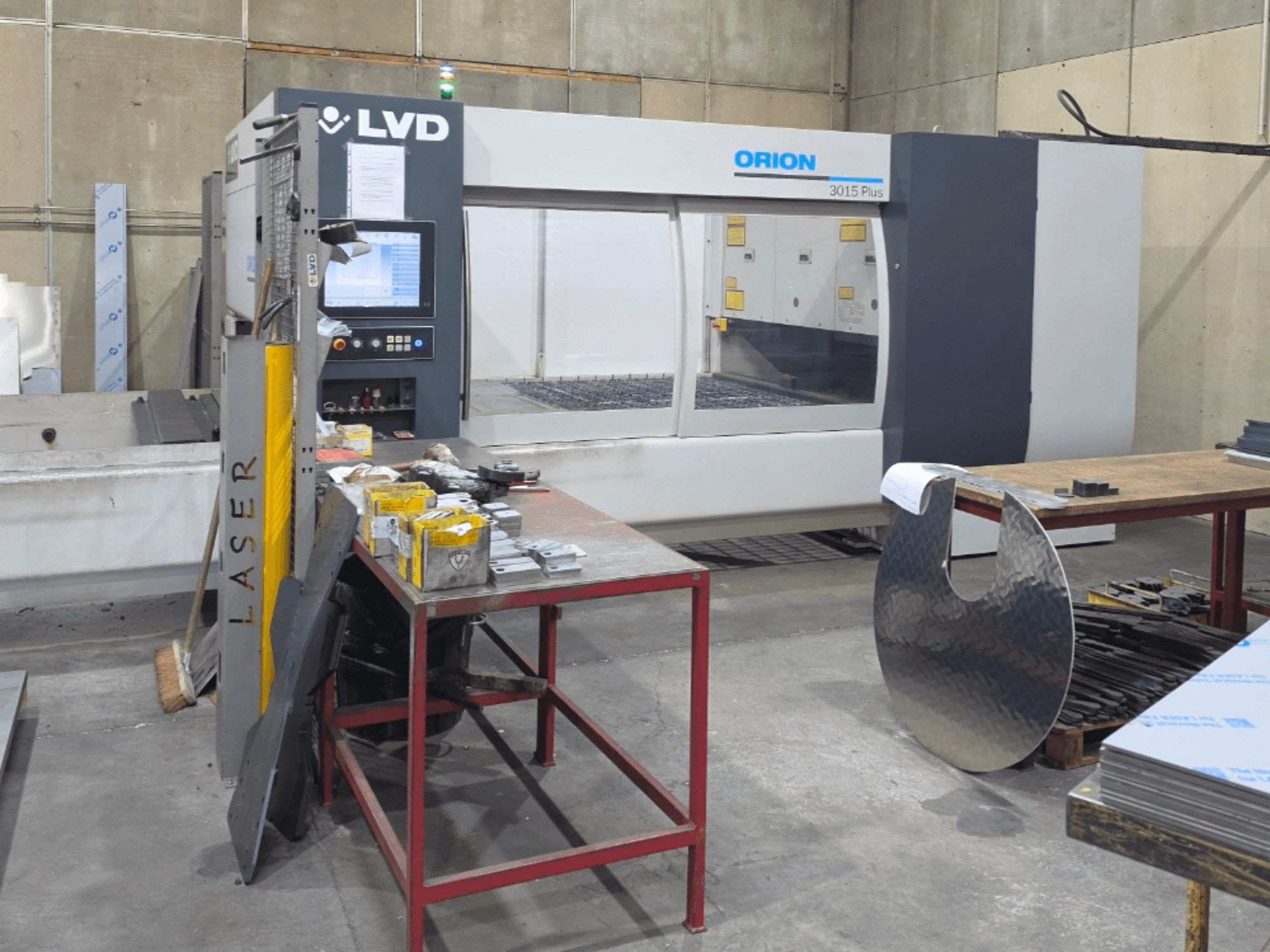 LVD Orion 3015 Plus CO₂-Laserschneidmaschine, Vorderansicht mit Bedienfeld; umgeben von Metallteilen auf einem Arbeitstisch.