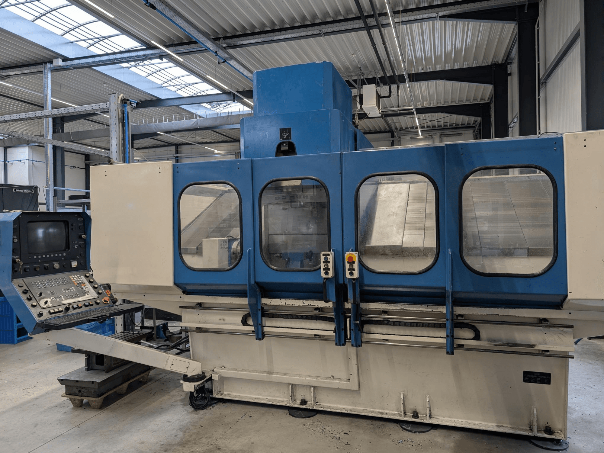 Blaue CNC-Industriemaschine mit Bedienfeld, Vorderansicht. Große Glasfenster, sichtbare Maschinenkomponenten und Tasten.