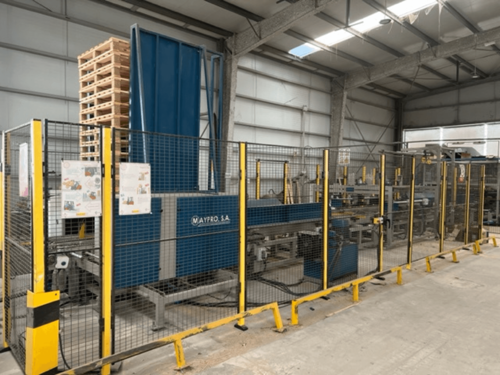 Industrielle Palettenverpackungsmaschine von Maypro S.A. in einem Lager, mit Schutzgitter und Bedienelementen, Seitenansicht.