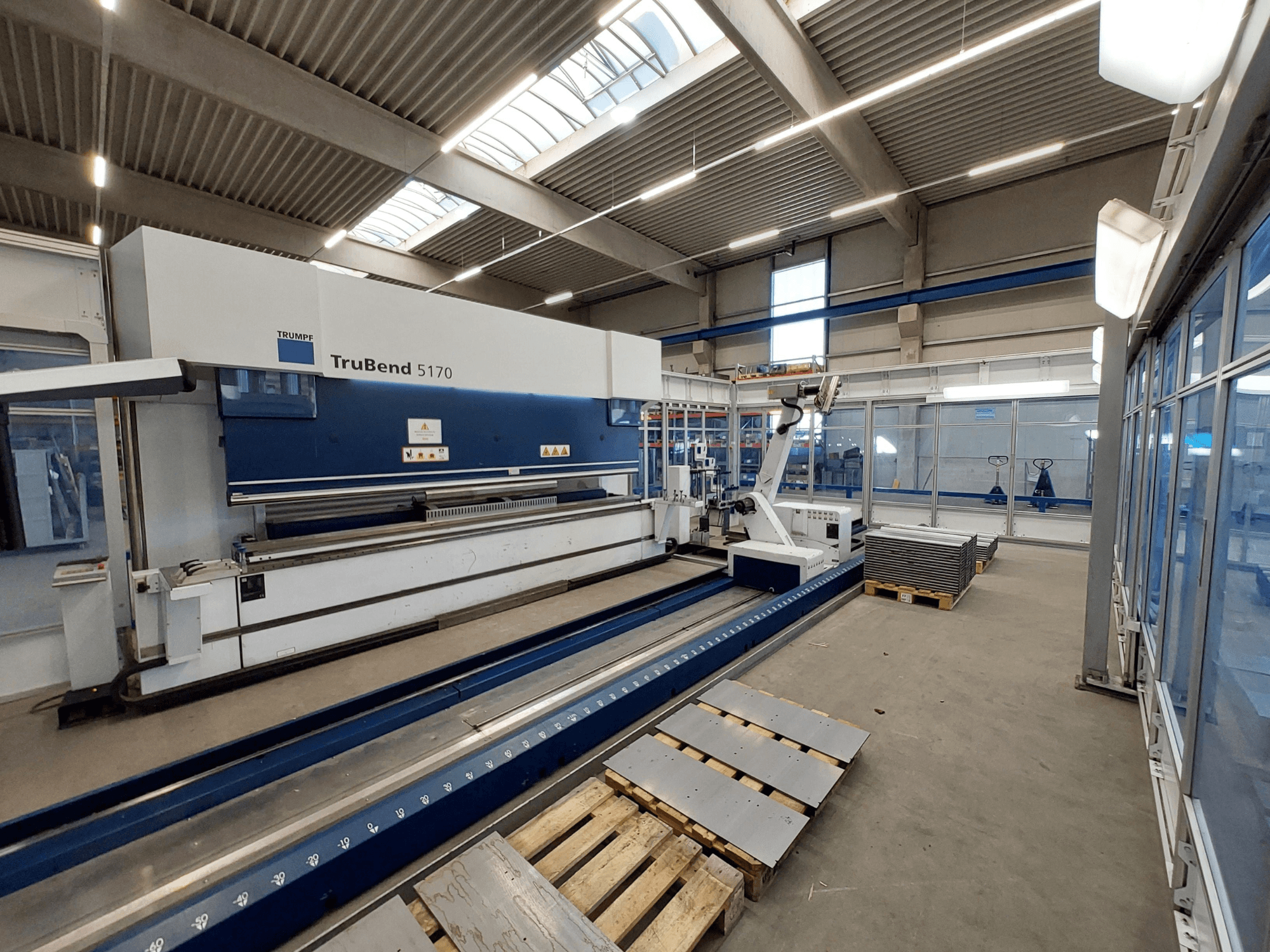 TRUMPF TruBend 5170 Abkantpresse in einer Werkstatt, Seitenansicht, mit Biegewerkzeug und gestapelten Blechen auf Paletten.