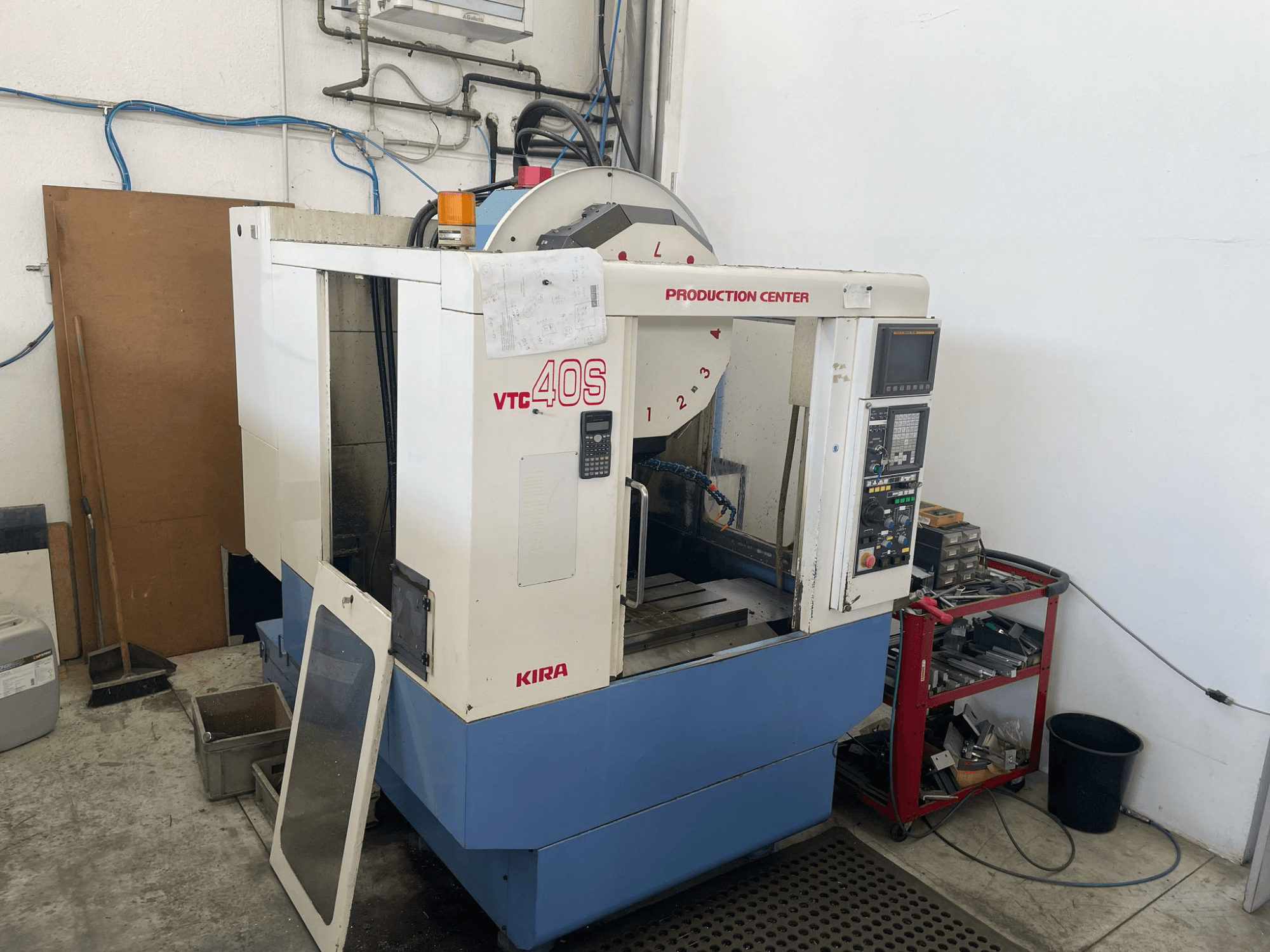KIRA VTC 40S Bearbeitungszentrum in der Seitenansicht, mit Bedienfeld, offener Tür und Werkzeugen auf einem Lagerwagen daneben.