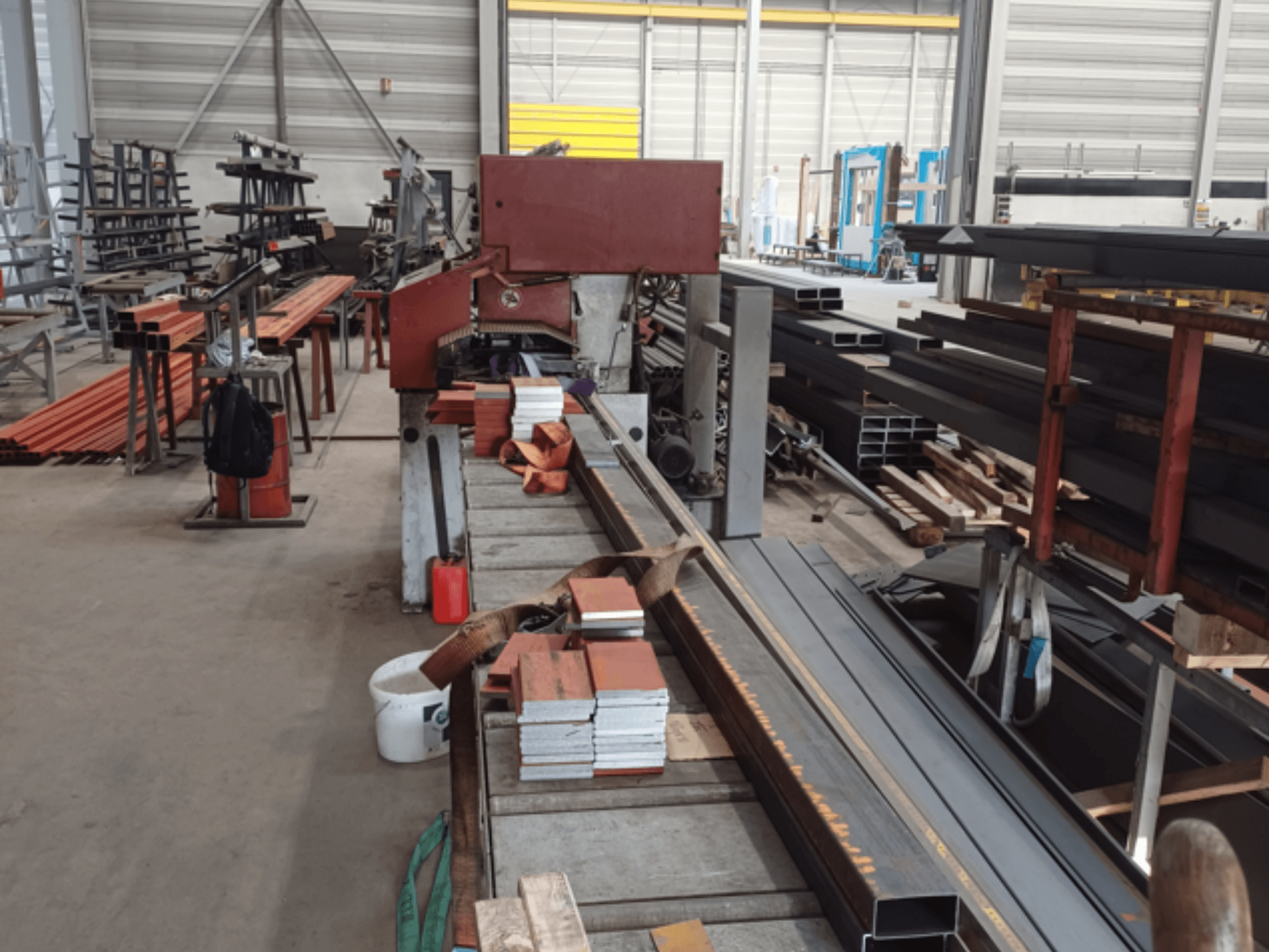 Fertigungswerkstatt mit verschiedenen Metallprofilen, mit einer roten Sägemaschine im Vordergrund und gestapelten Materialien.