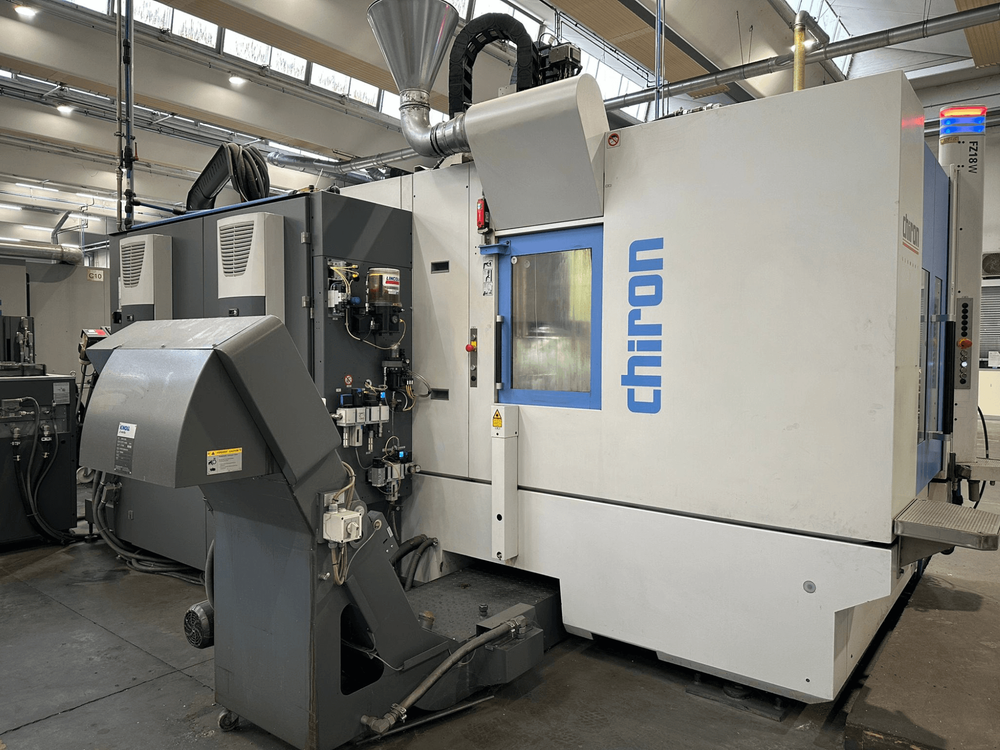 CNC-Maschine Chiron in Grau und Weiß, Vorderansicht, mit Bedienelementen, Rohren und einer Staubabsaugung.