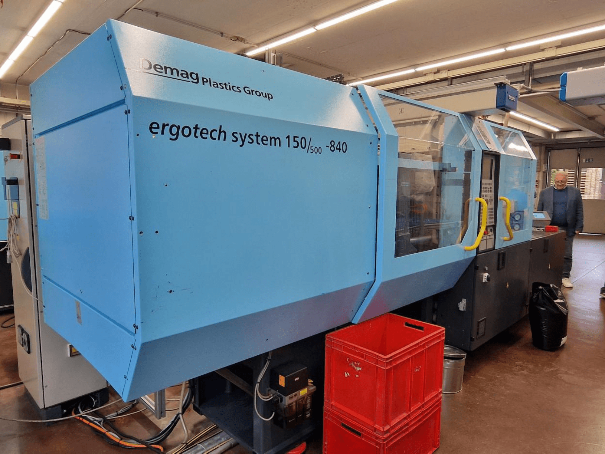 Demag Ergotech System 150/500-840 Spritzgießmaschine in blau, Seitenansicht, mit Kästen und Bediener im Hintergrund.