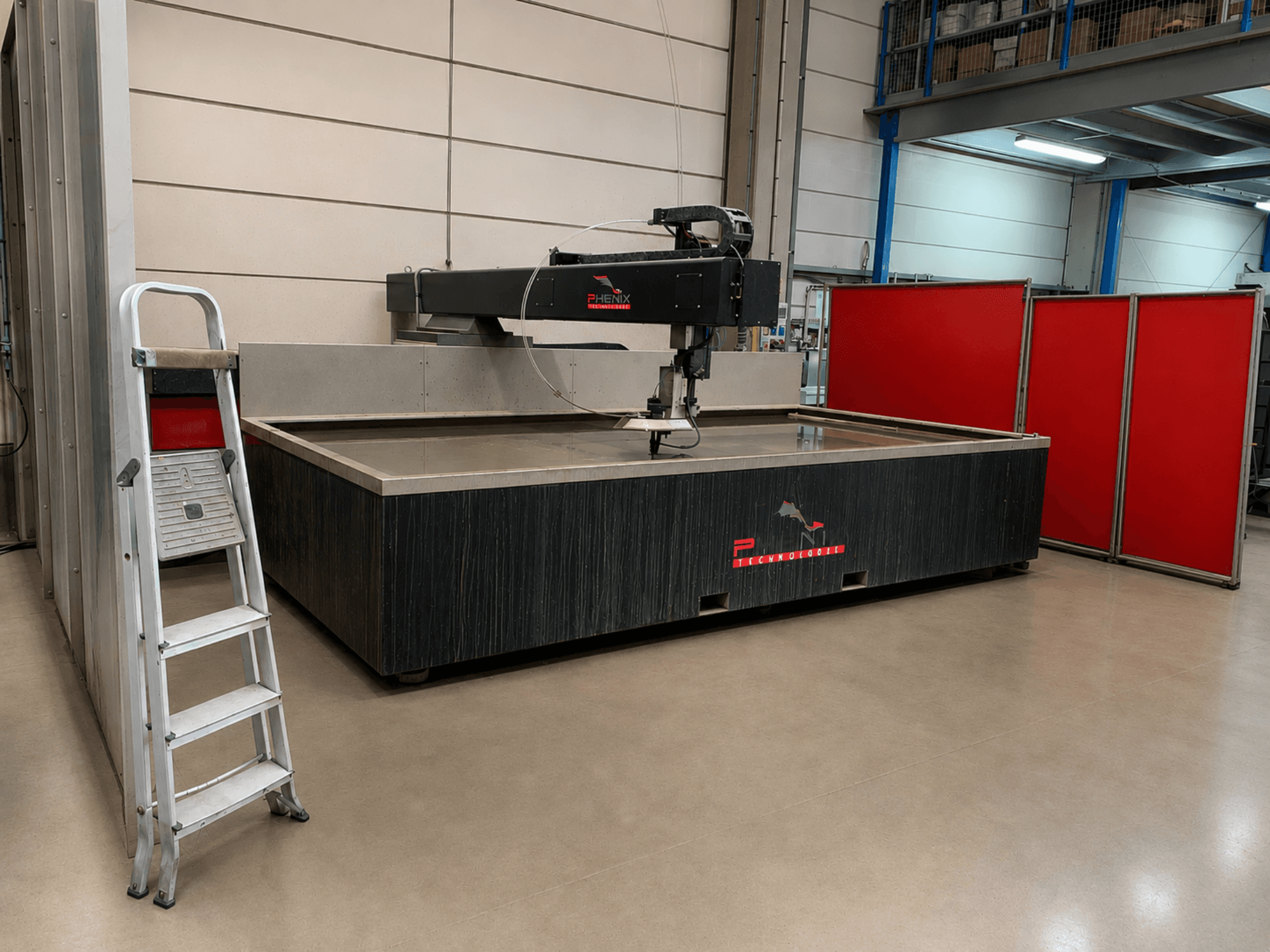 Wasserstrahlschneidemaschine PHENIX TECHNOLOGIE MD-JET 2040 in Seitenansicht, daneben eine silberne Leiter und rote Trennwände.