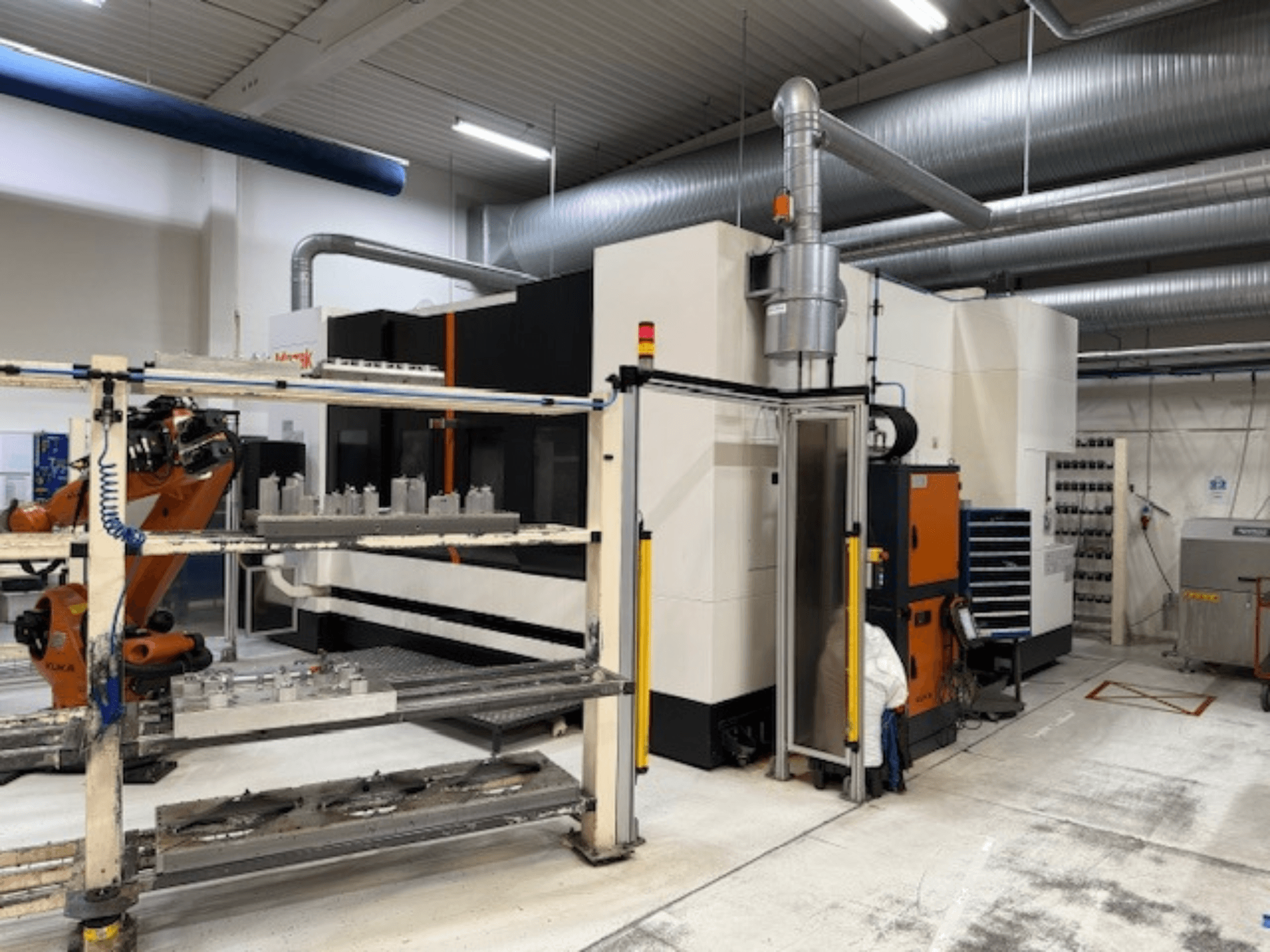Industrielle CNC-Maschine mit Roboterarm und Sicherheitsschranken, mit einer Frontansicht der Einrichtung in einem luftigen Arbeitsraum.