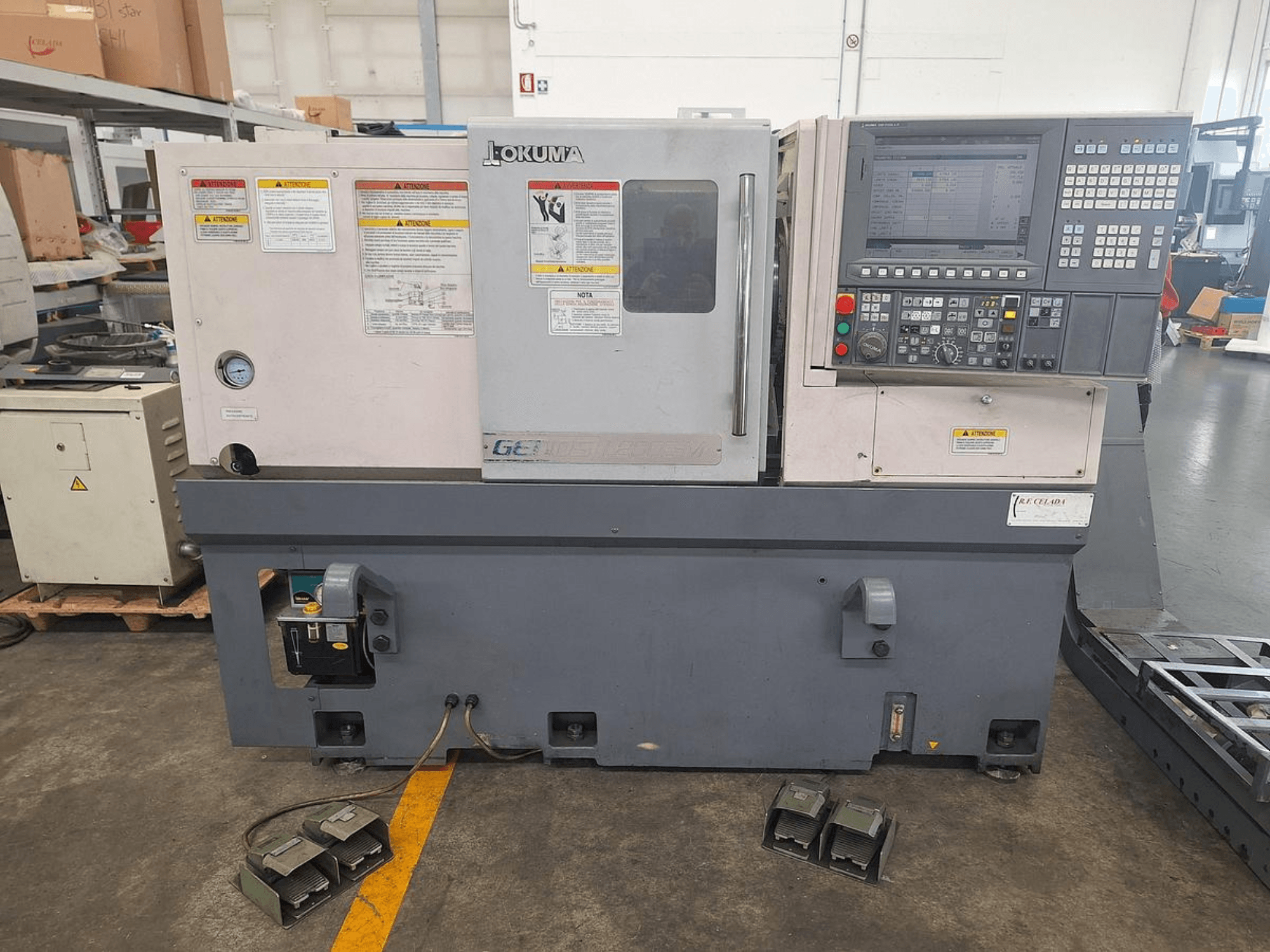 Okuma GENOS L200E-M Horizontaldrehmaschine in der Seitenansicht; mit Bedienfeld, Sicherheitswarnungen und Fußpedalen.